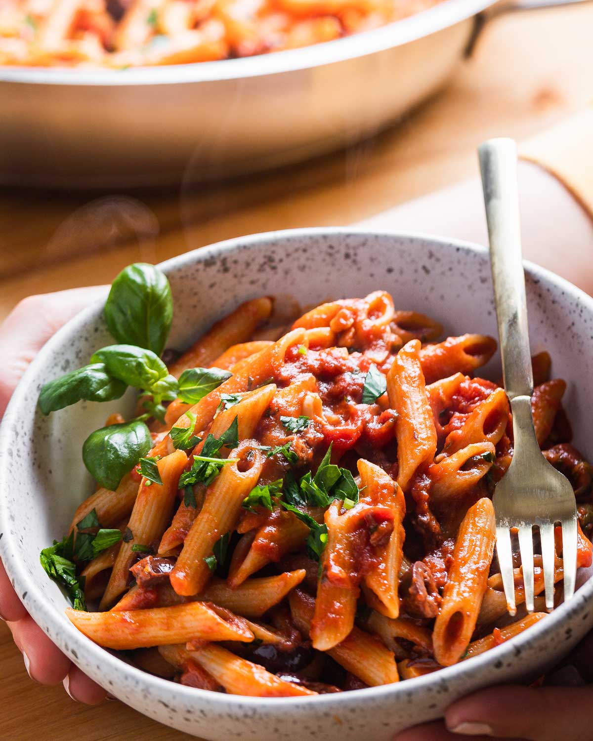 Hands holding bowl of penne puttanesca.