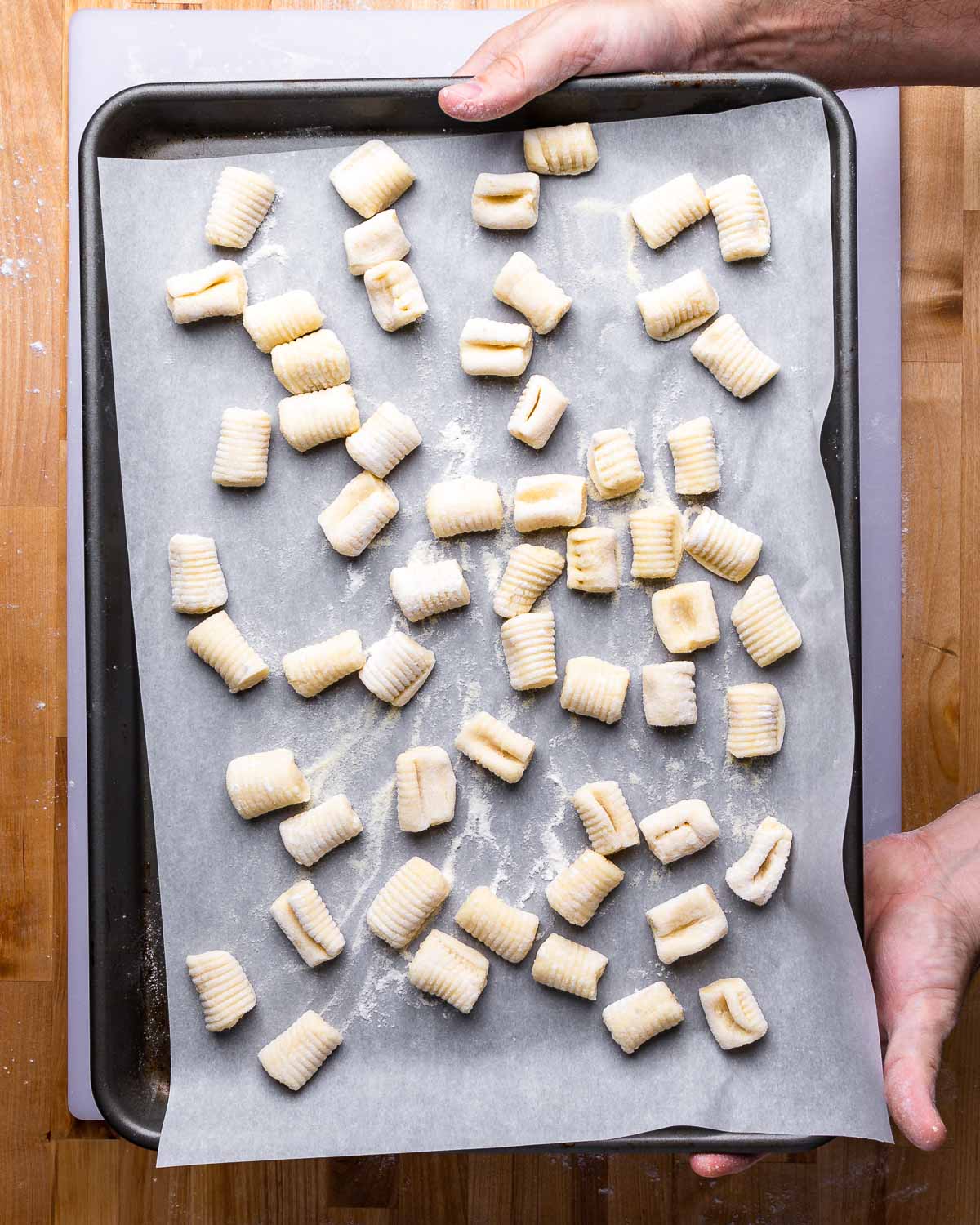 Hands holding parchment paper lined tray of uncooked potato gnocchi.