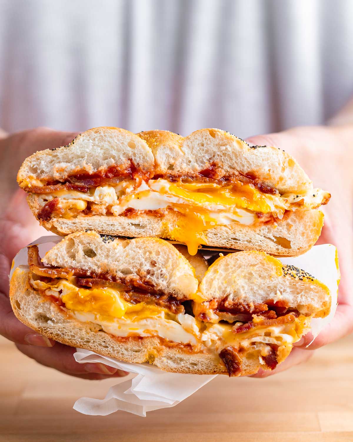 Hands holding a bacon egg and cheese sandwich cut in half with the yolk dripping.