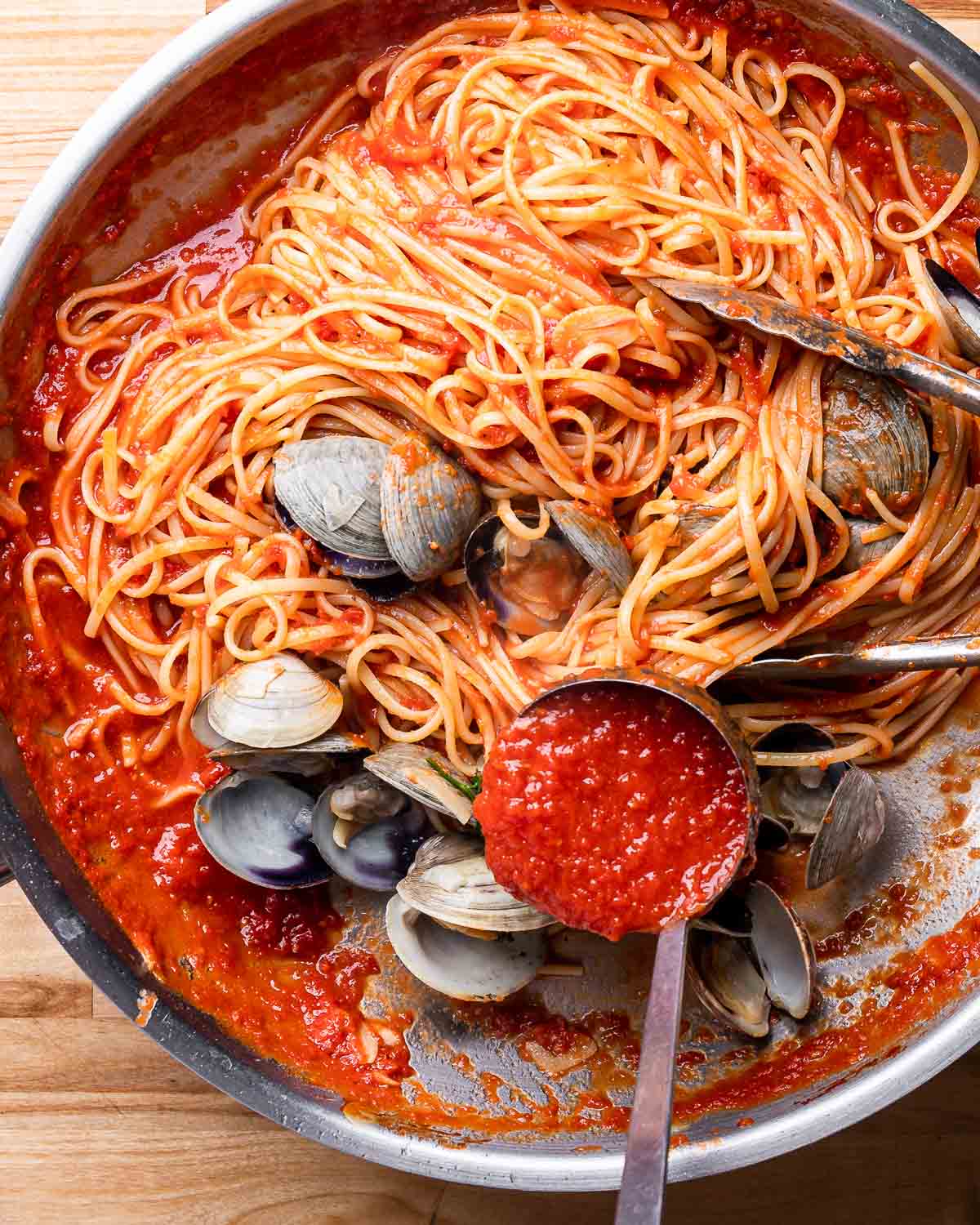 Large pan with linguine and clams with ladle of red sauce being poured on top.