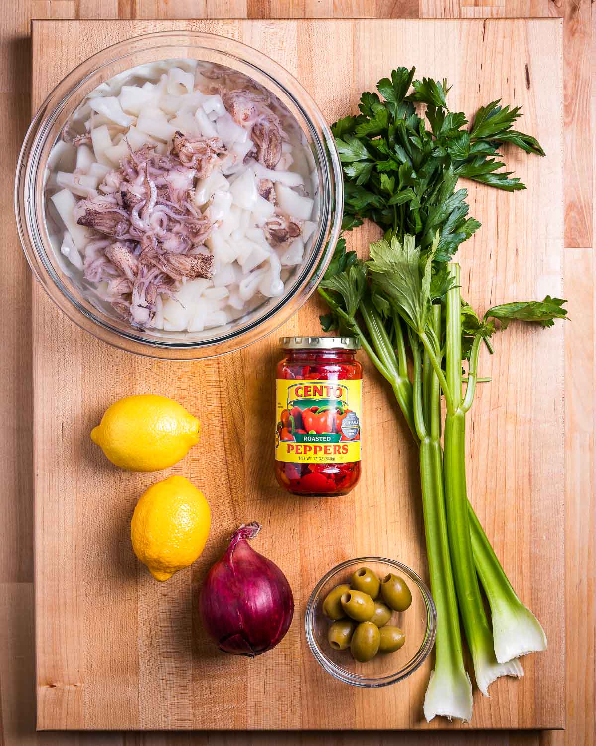 Ingredients shown: bowl of calamari, celery, parsley, lemons, roasted red peppers, red onion, and olives.