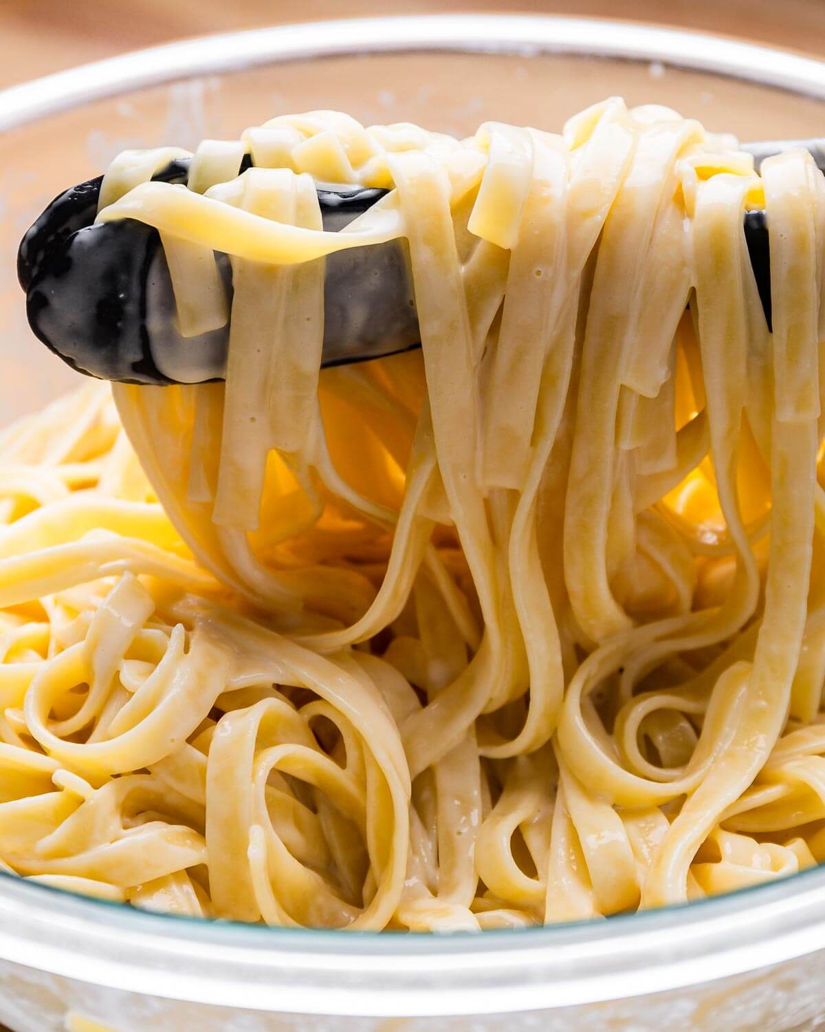 Tongs holding cooked fettuccine alfredo in large glass bowl.