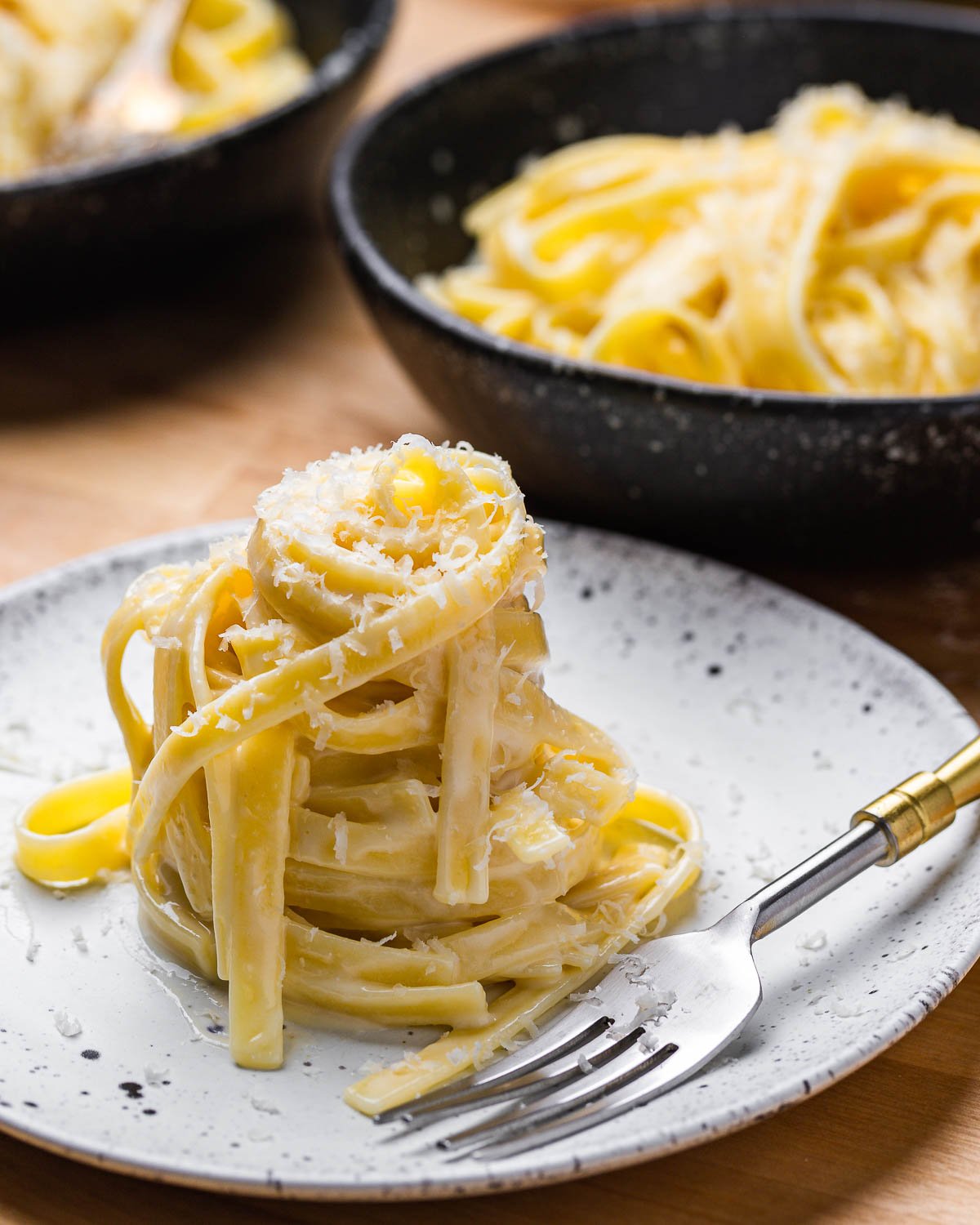 White plate with nest of fettuccine alfredo along with 2 black bowls in background.