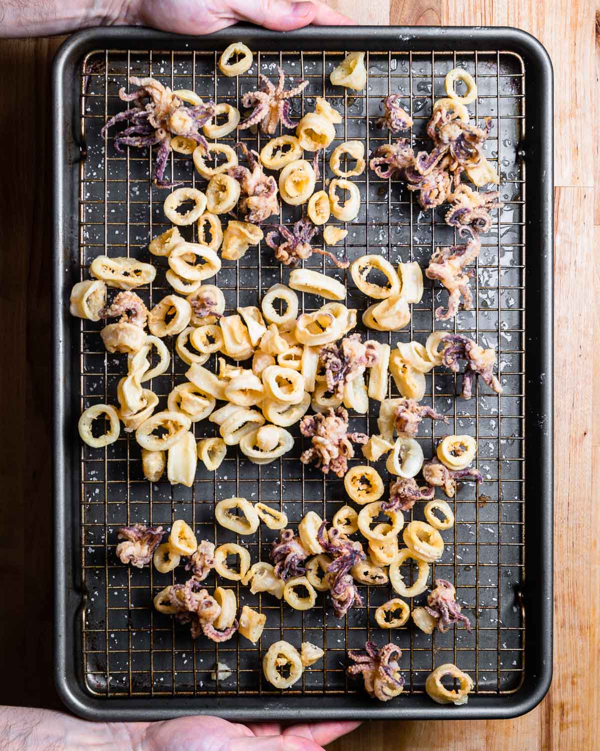 Hands holding baking sheet and wire rack with fried calamari.