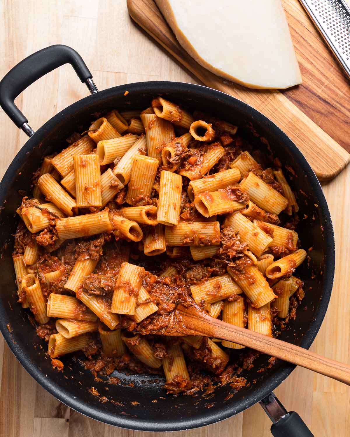 Large black pan with rigatoni lamb ragu and block of Parmigiano Reggiano on the side.