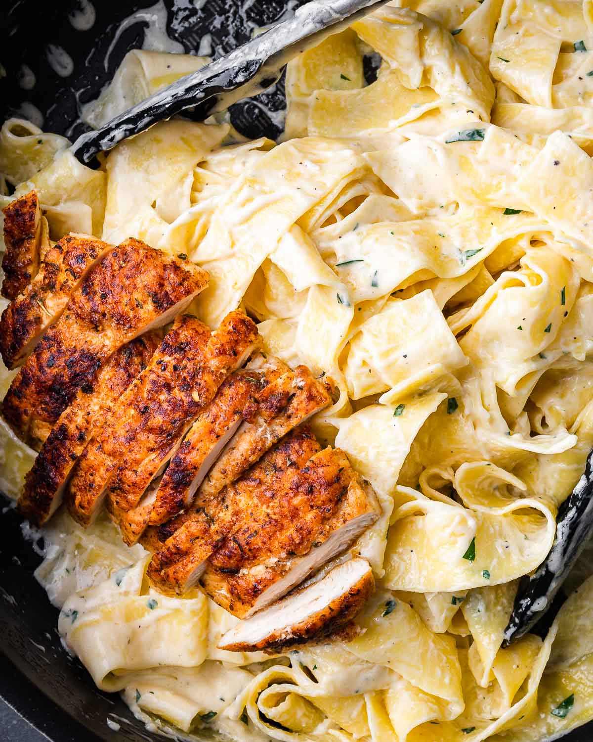 Chicken alfredo pasta in black pan with tongs.