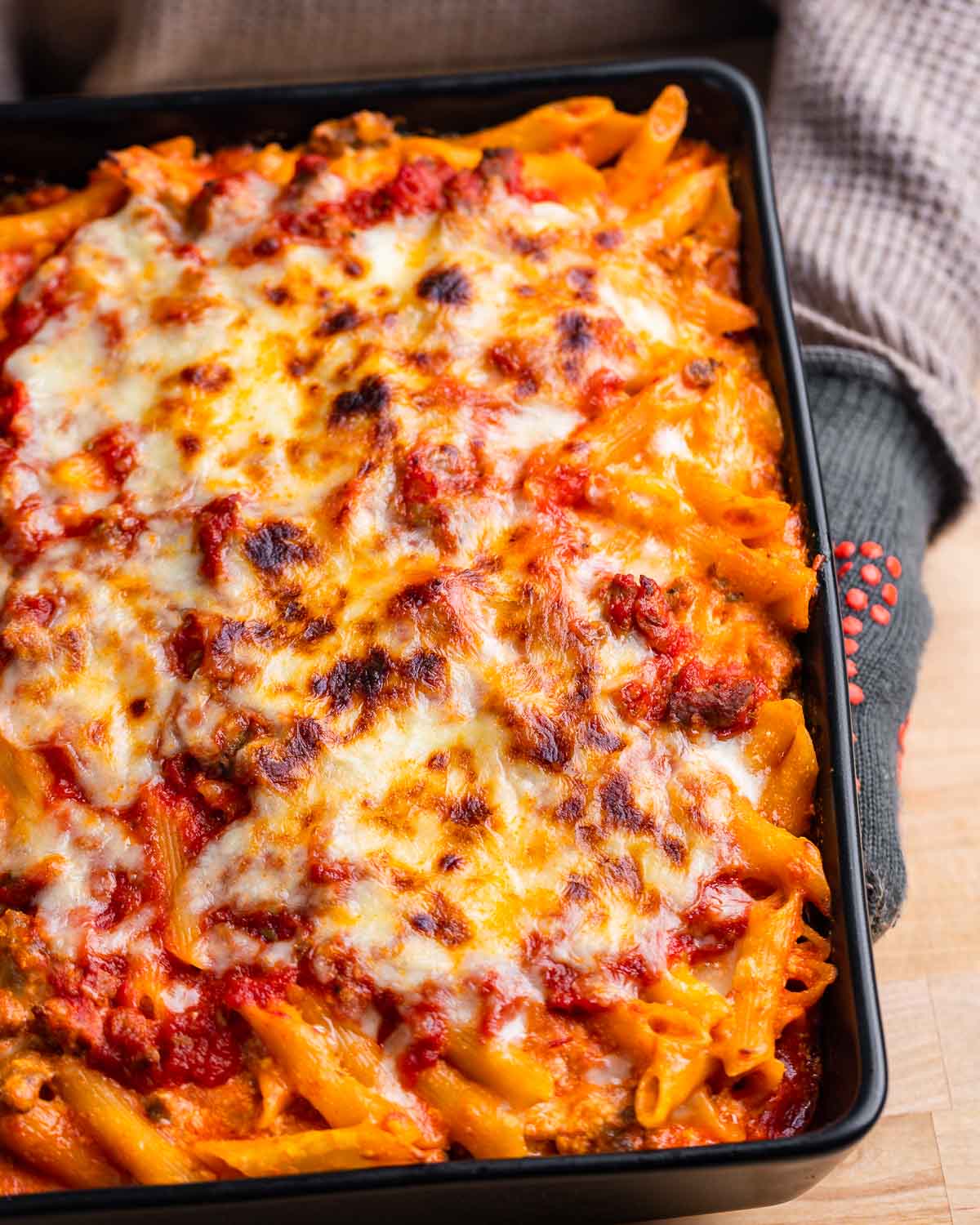 Gloves holding baked penne pasta in black baking dish.