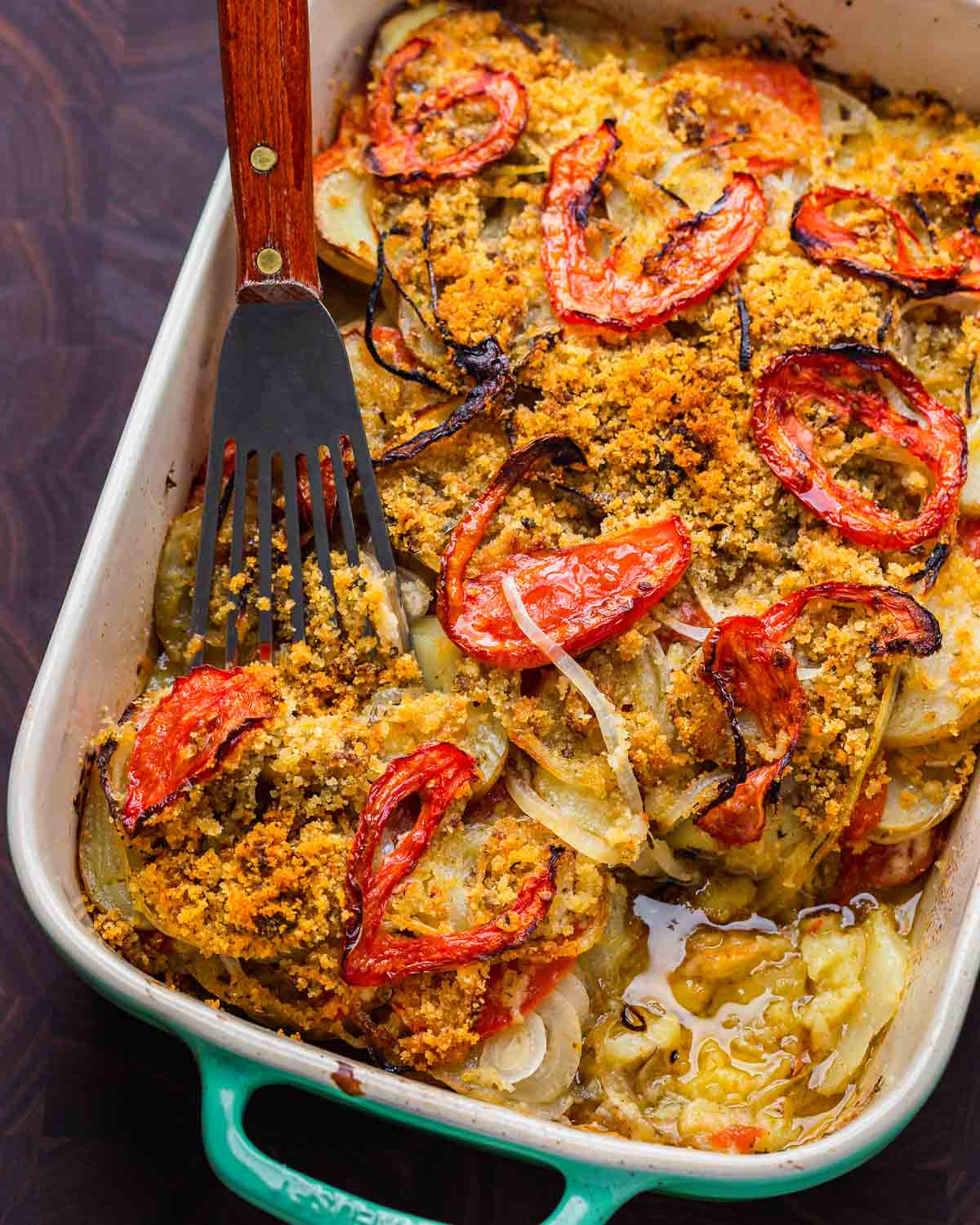 Italian potato casserole with spatula in baking dish on wooden cutting board.