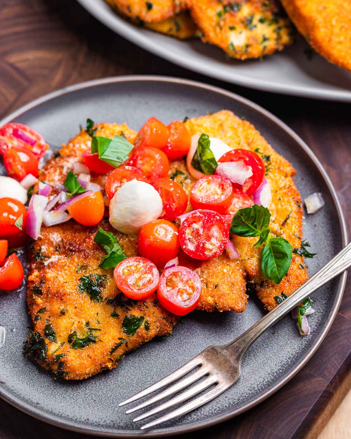 Grey plate with large chicken cutlet topped with tomato and mozzarella balls.