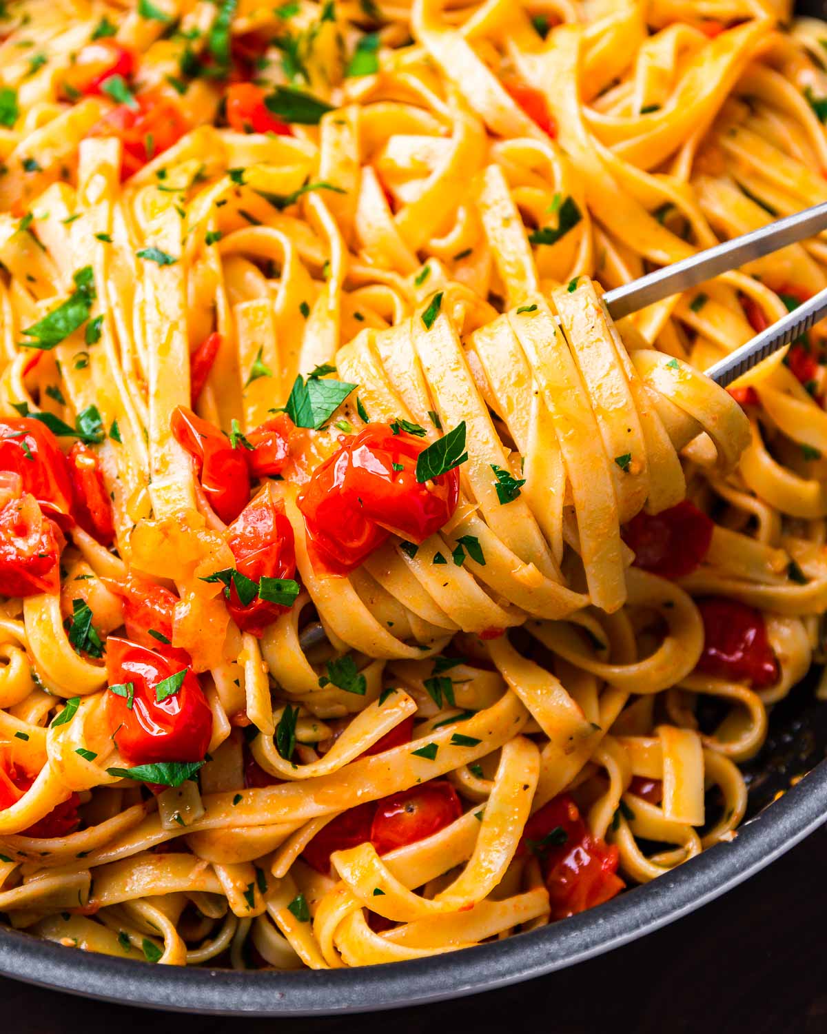 Kitchen tweezers holding fettuccine with cherry tomato butter sauce in black pan.