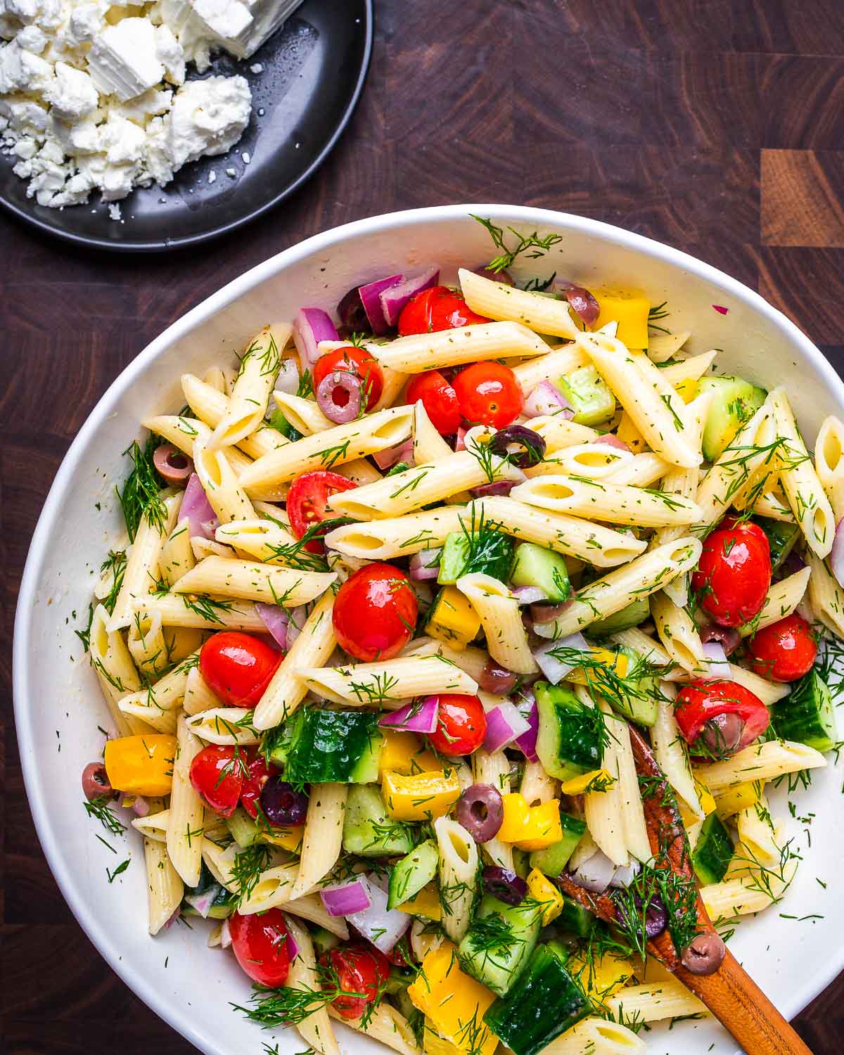 Greek pasta salad in large serving bowl with plate of feta cheese on the side.