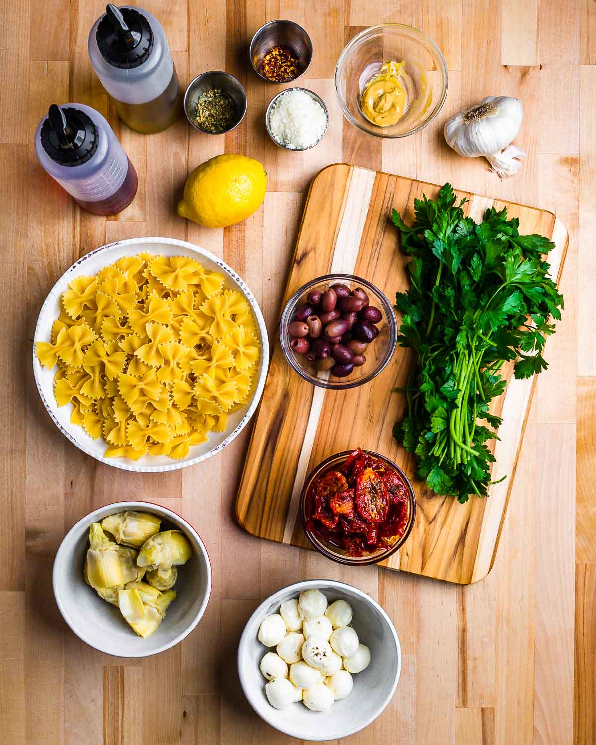 Ingredients shown: oil, vinegar, lemons, cheese, mustard, garlic, bow tie pasta, olives, sun dried tomatoes, artichoke hearts, and parsley.