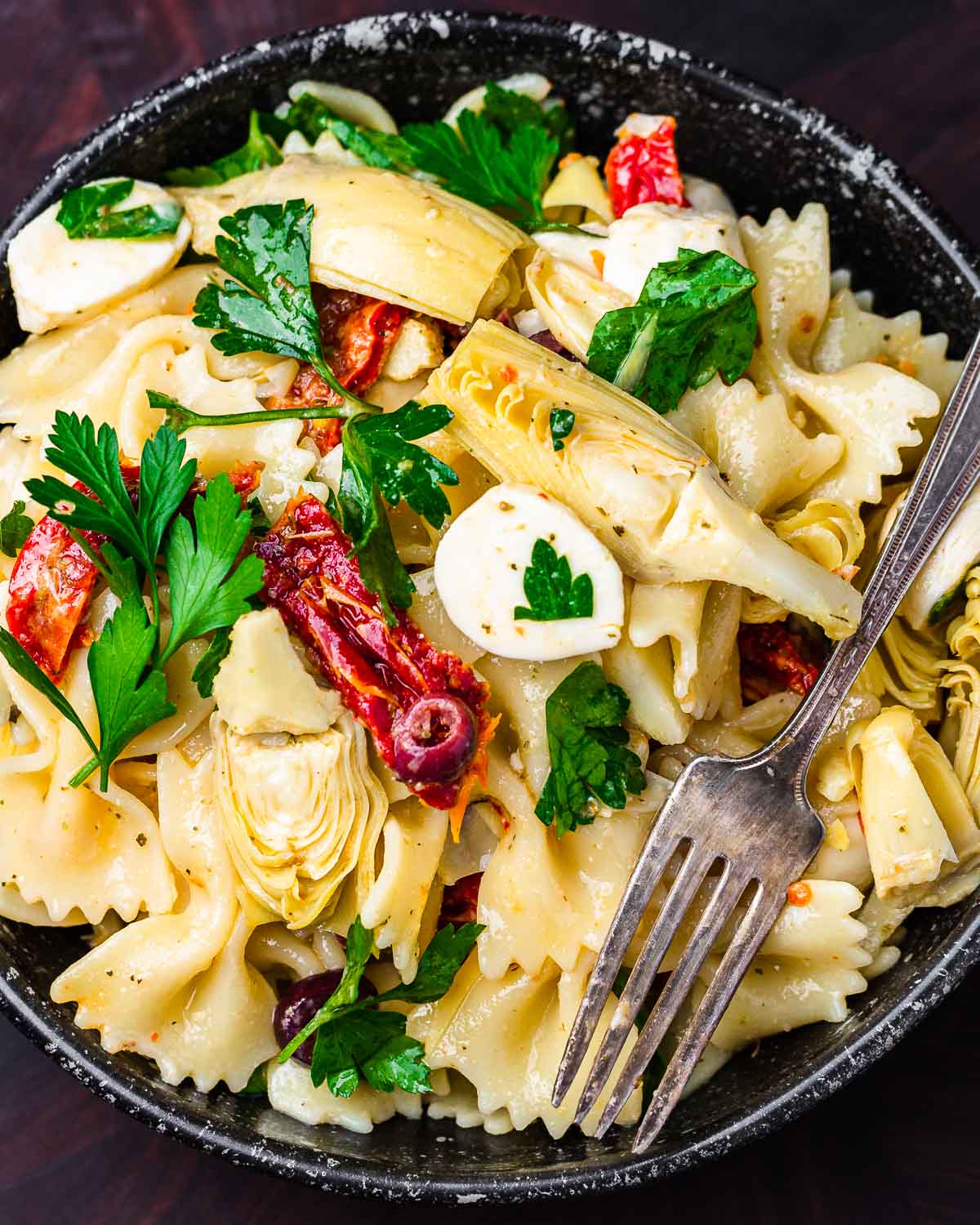 Pasta salad with artichokes and sun dried tomatoes in black bowl.