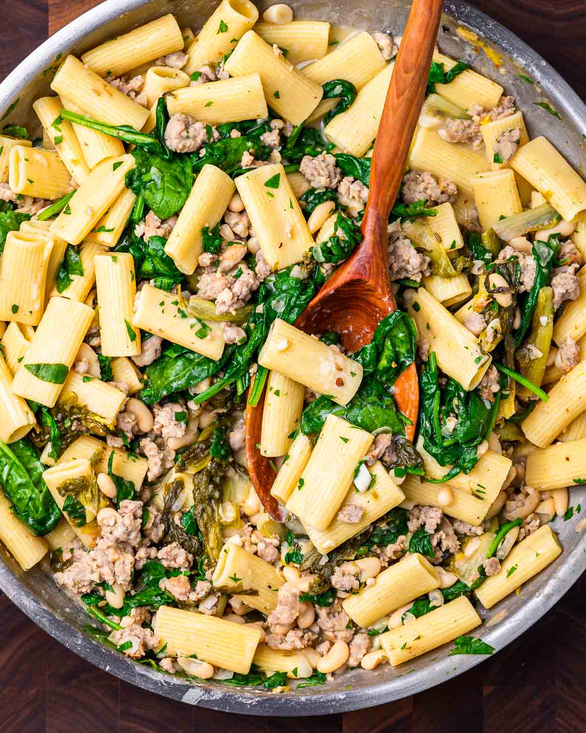Overhead shot of beans and greens sausage pasta in large pan with wooden ladle.