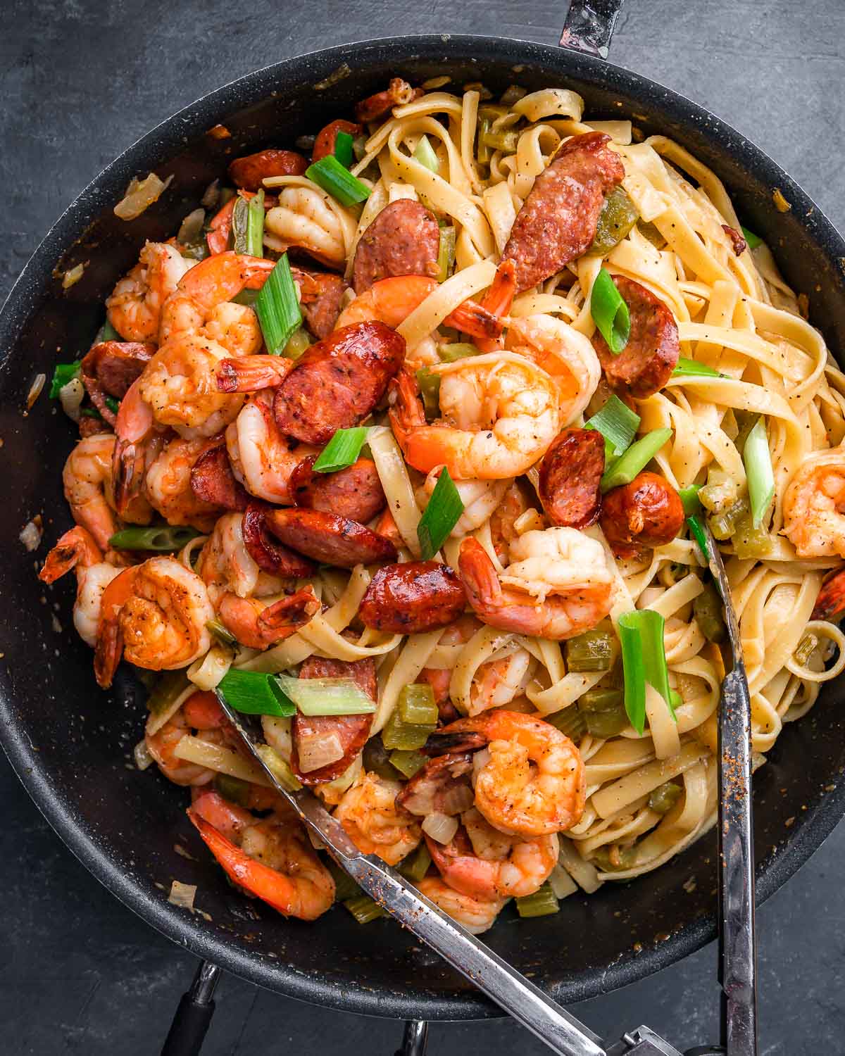 Overhead shot of Cajun shrimp and sausage pasta in large pan with tongs.