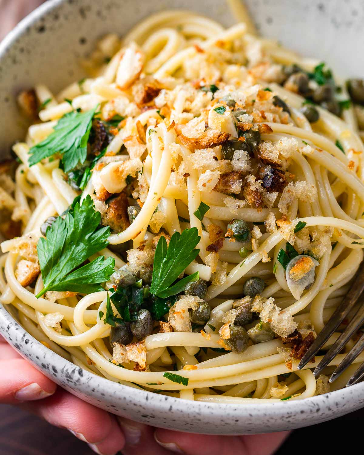 Hands holding lemon caper pasta in white bowl.