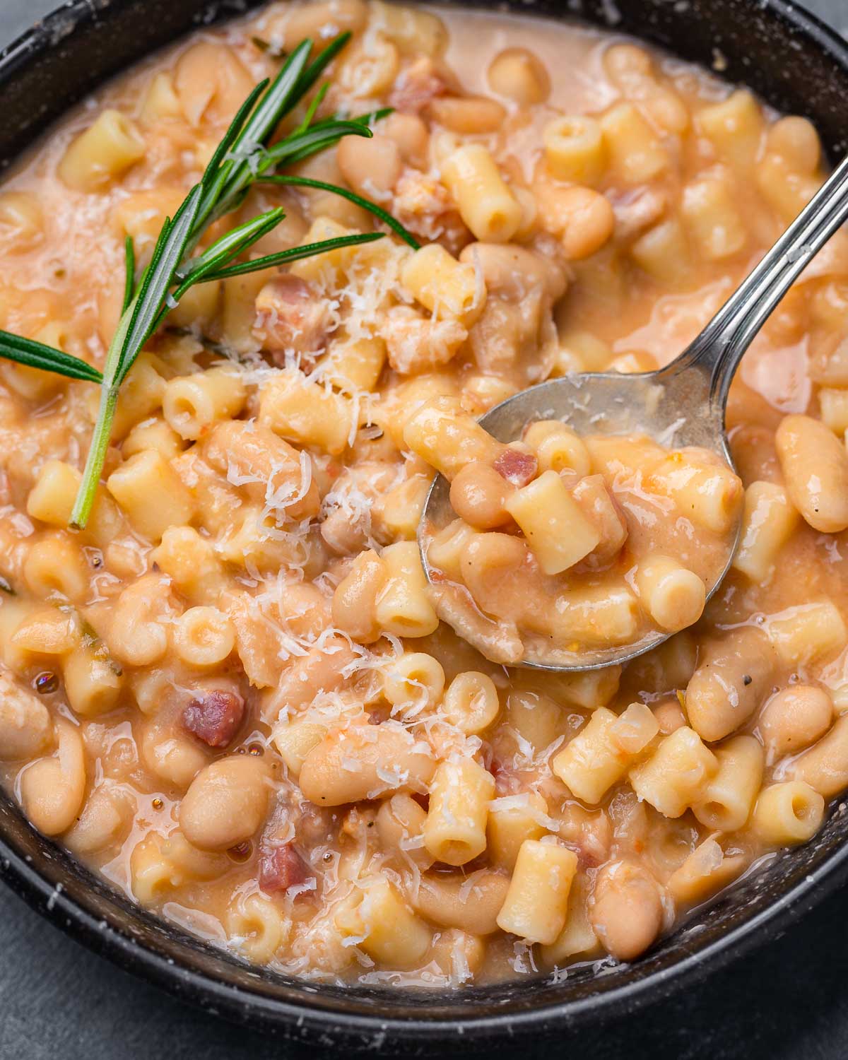 Pasta e fagioli in black bowl with spoon and rosemary sprig garnish.