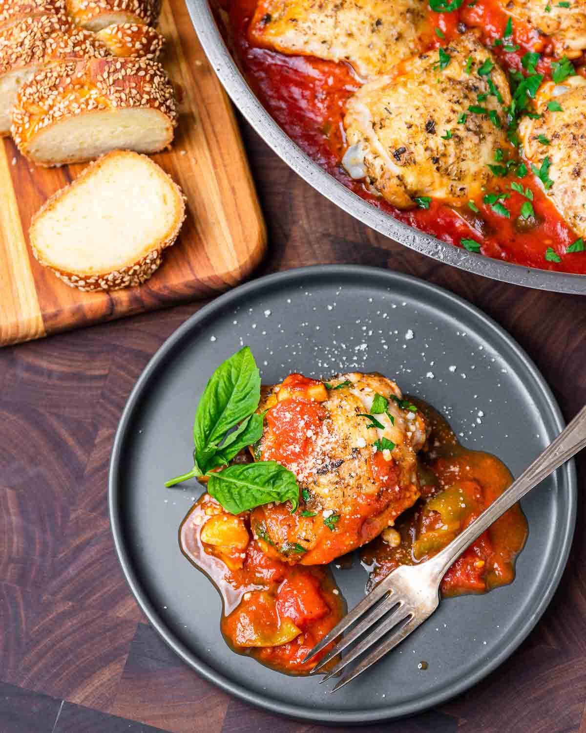 Grey plate with chicken fra diavolo along with cut loaf of breaf and large pan.