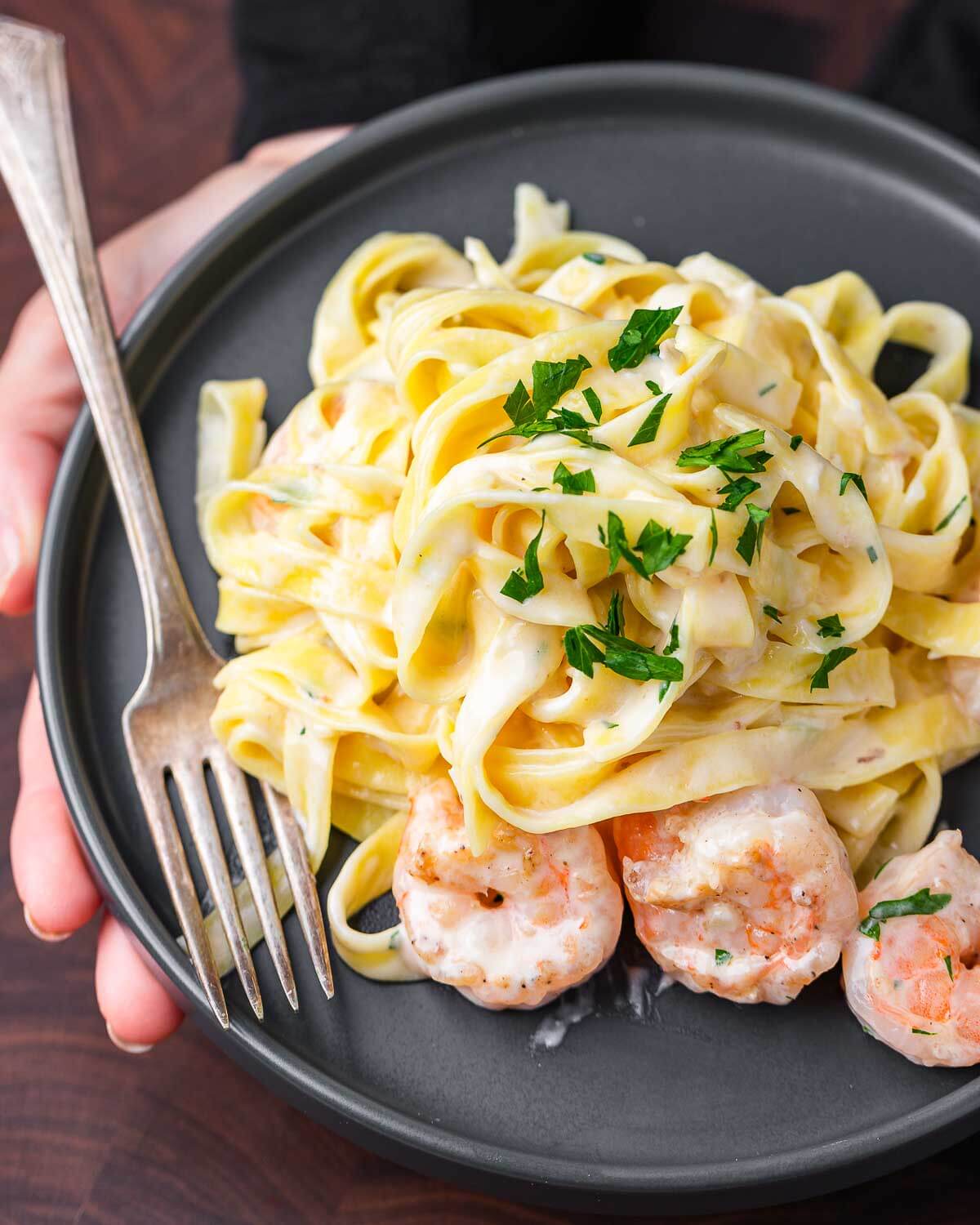 Hands holding shrimp alfredo in grey plate.