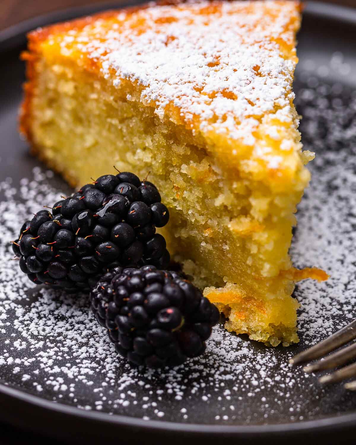 Piece of orange olive cake in plate with powdered sugar and blackberries.