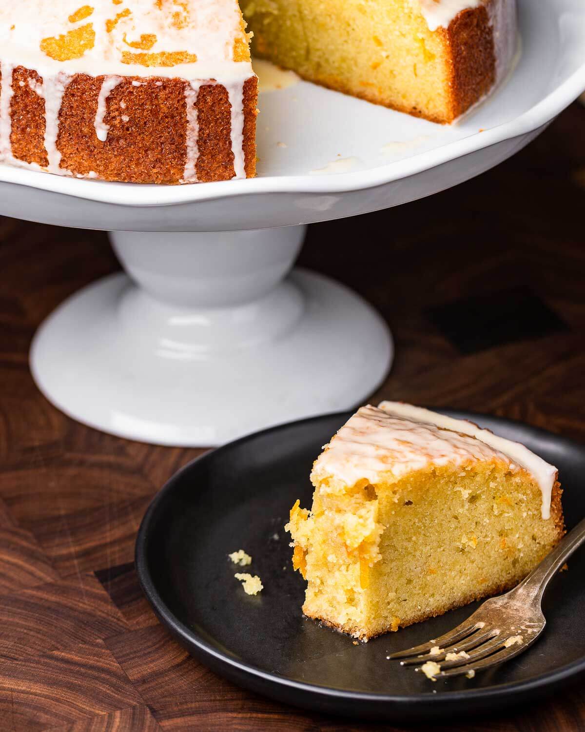 Cut piece of orange olive cake on black plate with cake stand in background.