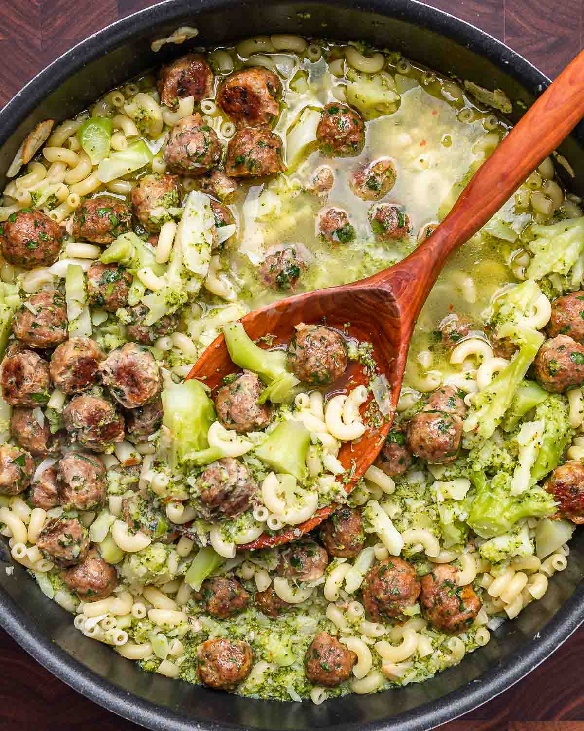 Overhead shot of large pan with pasta broccoli and tiny meatballs with large wooden spoon.