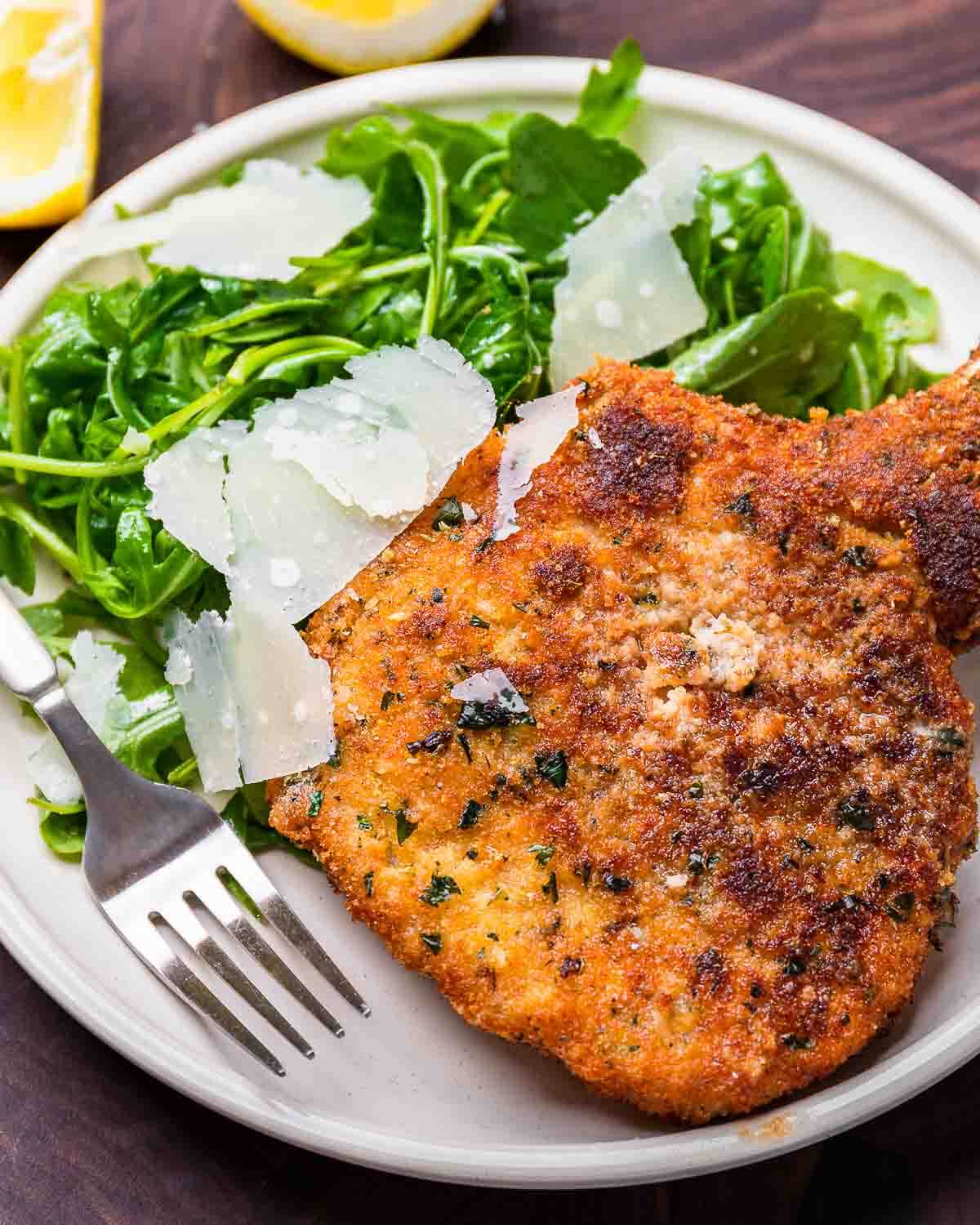 Pork chop Milanese with arugula salad and shaved parmesan on white plate.