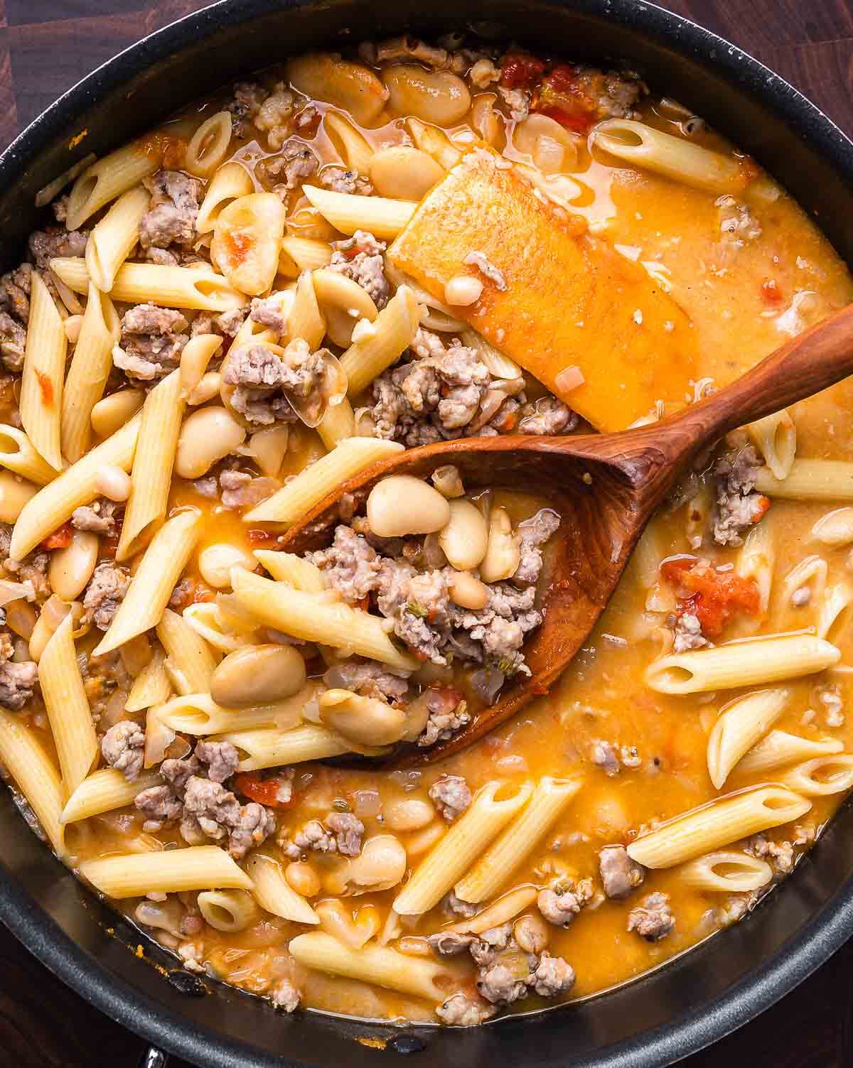 Overhead shot of sausage pasta fagioli in black pot with wooden spoon.