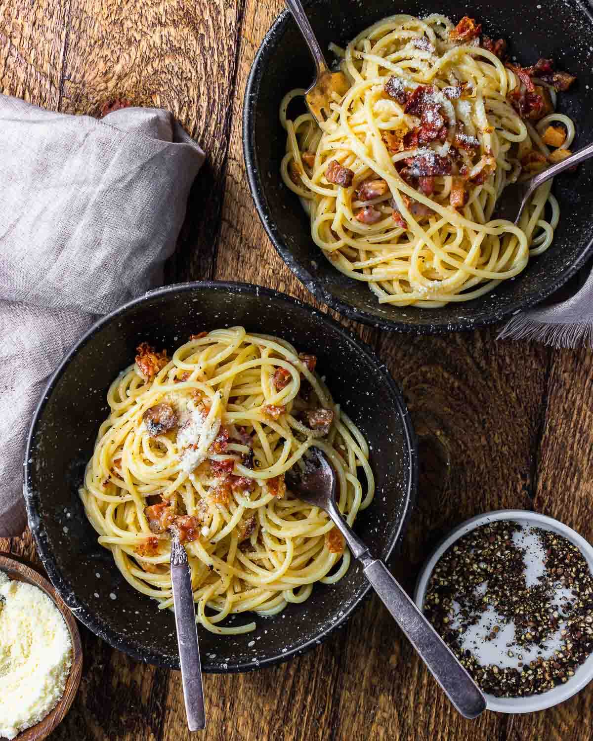 Overhead shot of two bowls of spaghetti carbonara with small bowl of pepper and pecorino.