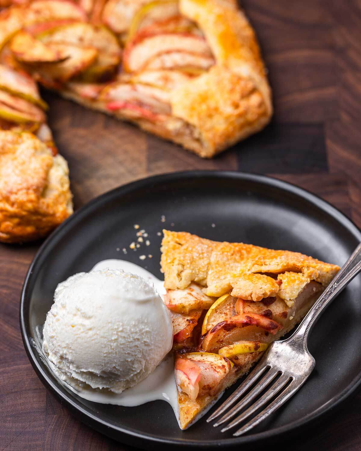 Slice of apple crostata and vanilla ice cream in black plate on walnut cutting board.