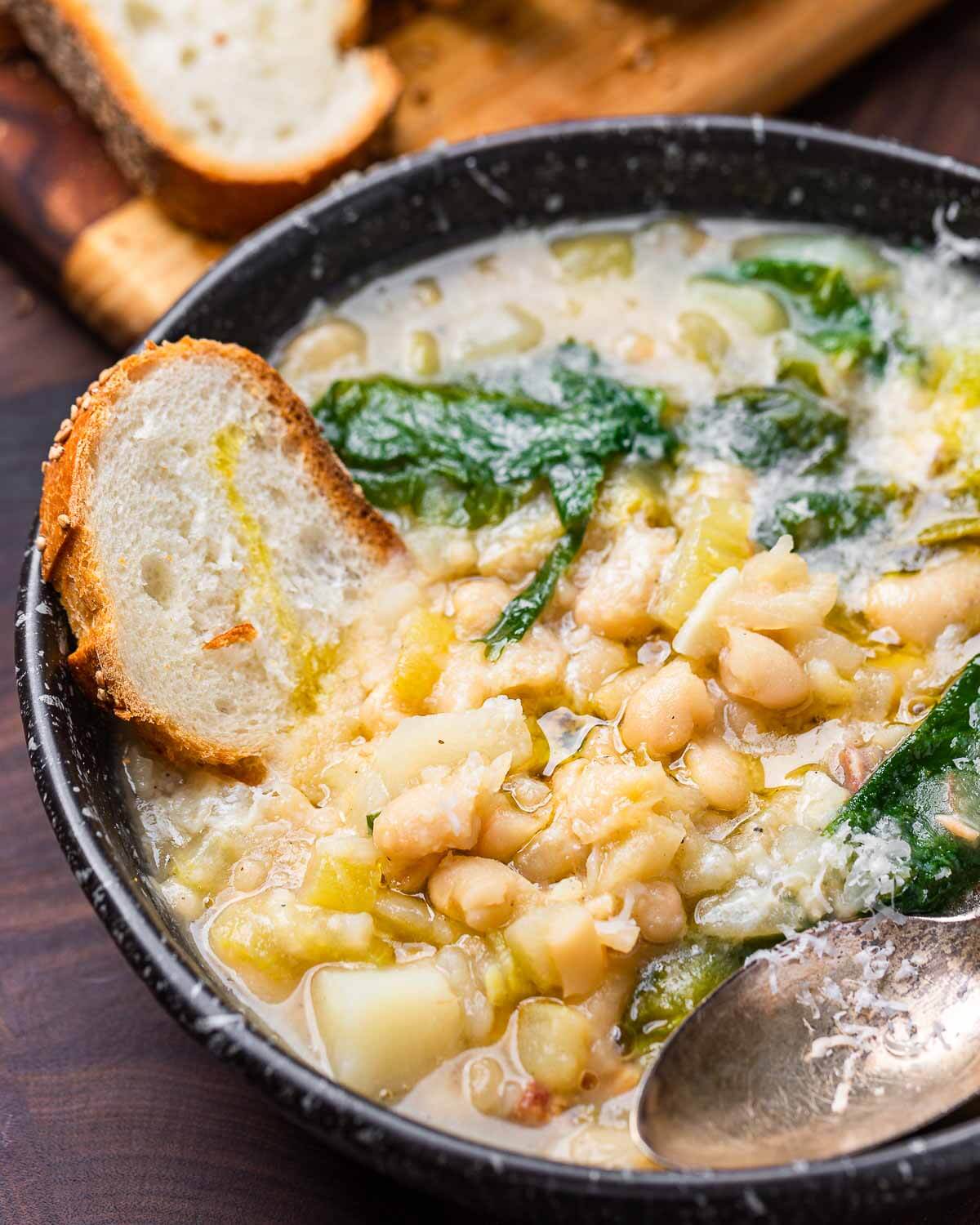 Escarole bean soup in black bowl with piece of bread dipped into bowl.