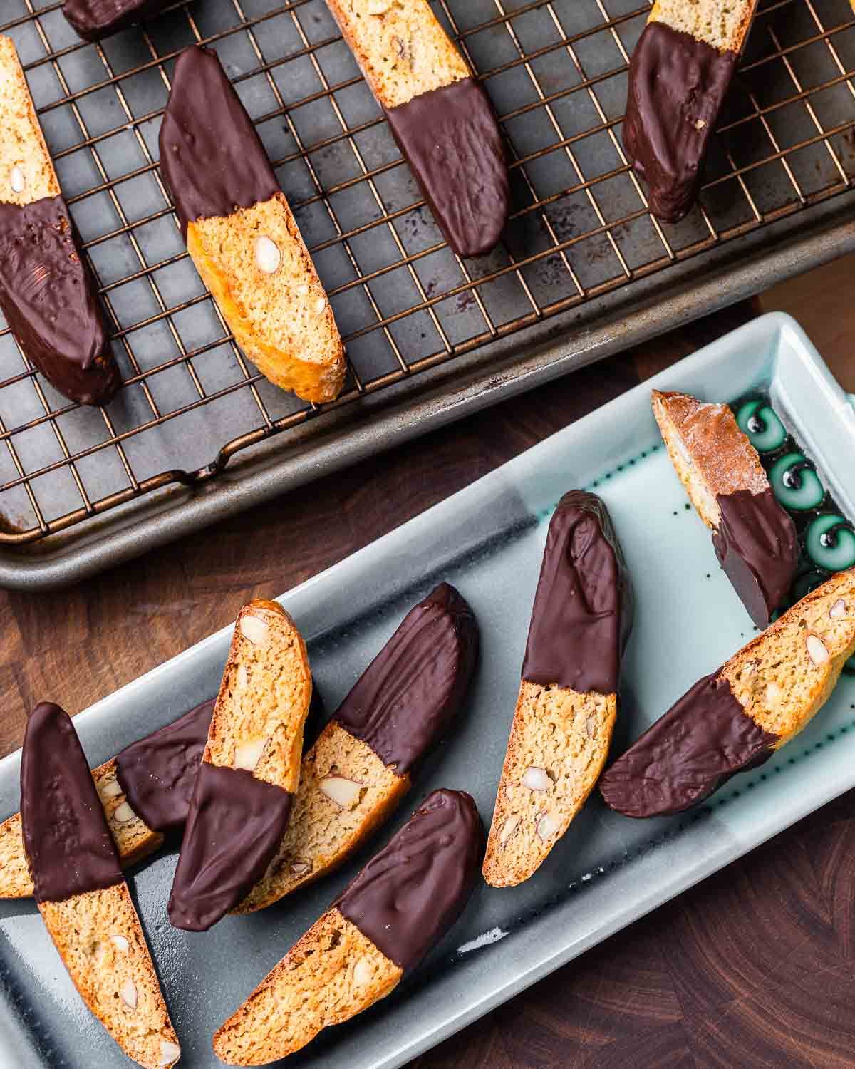 Almond biscotti dipped in chocolate on blue plate along with more biscotti on wire rack in the background.