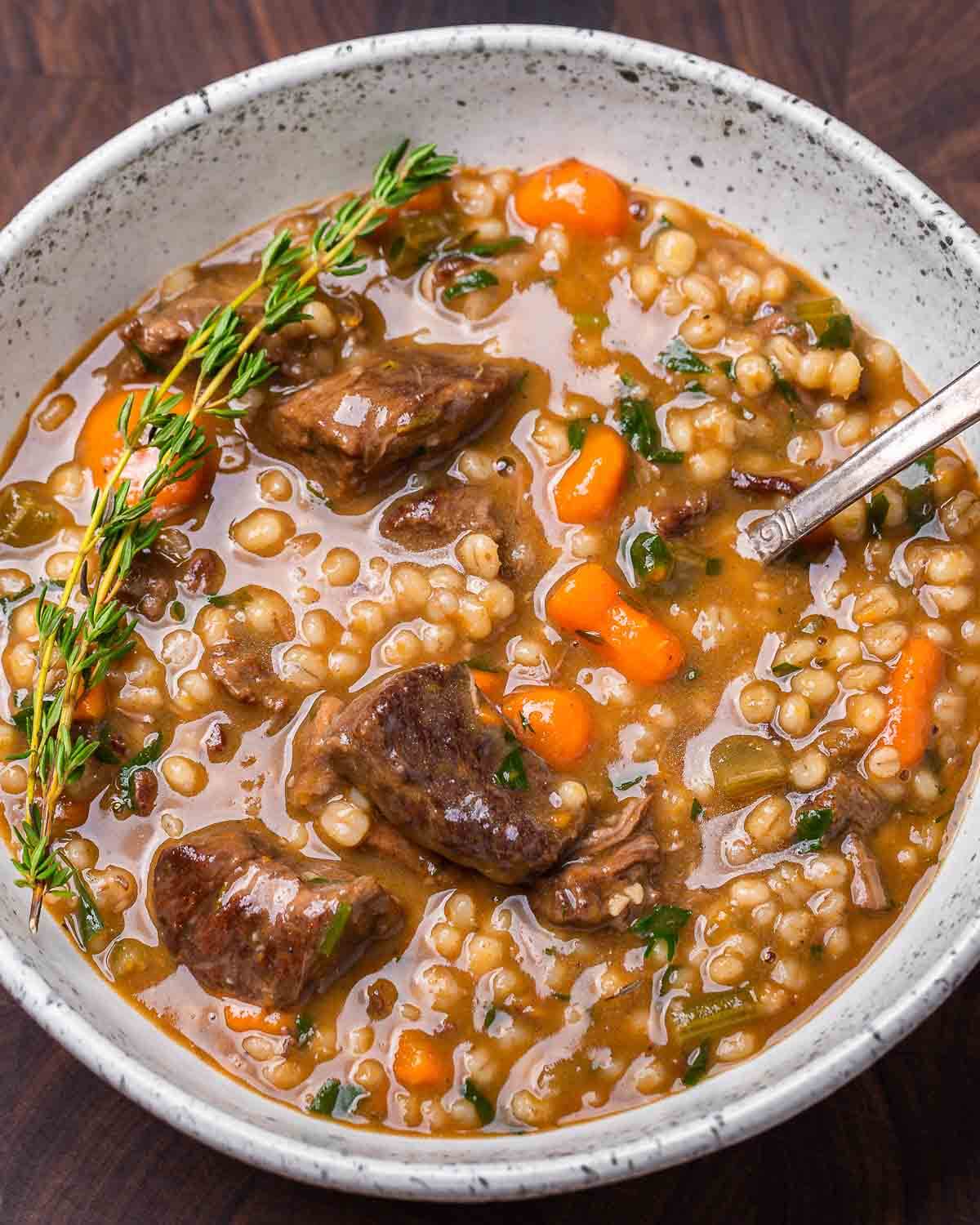 Overhead shot of beef barley soup in white plate with a sprig of thyme.