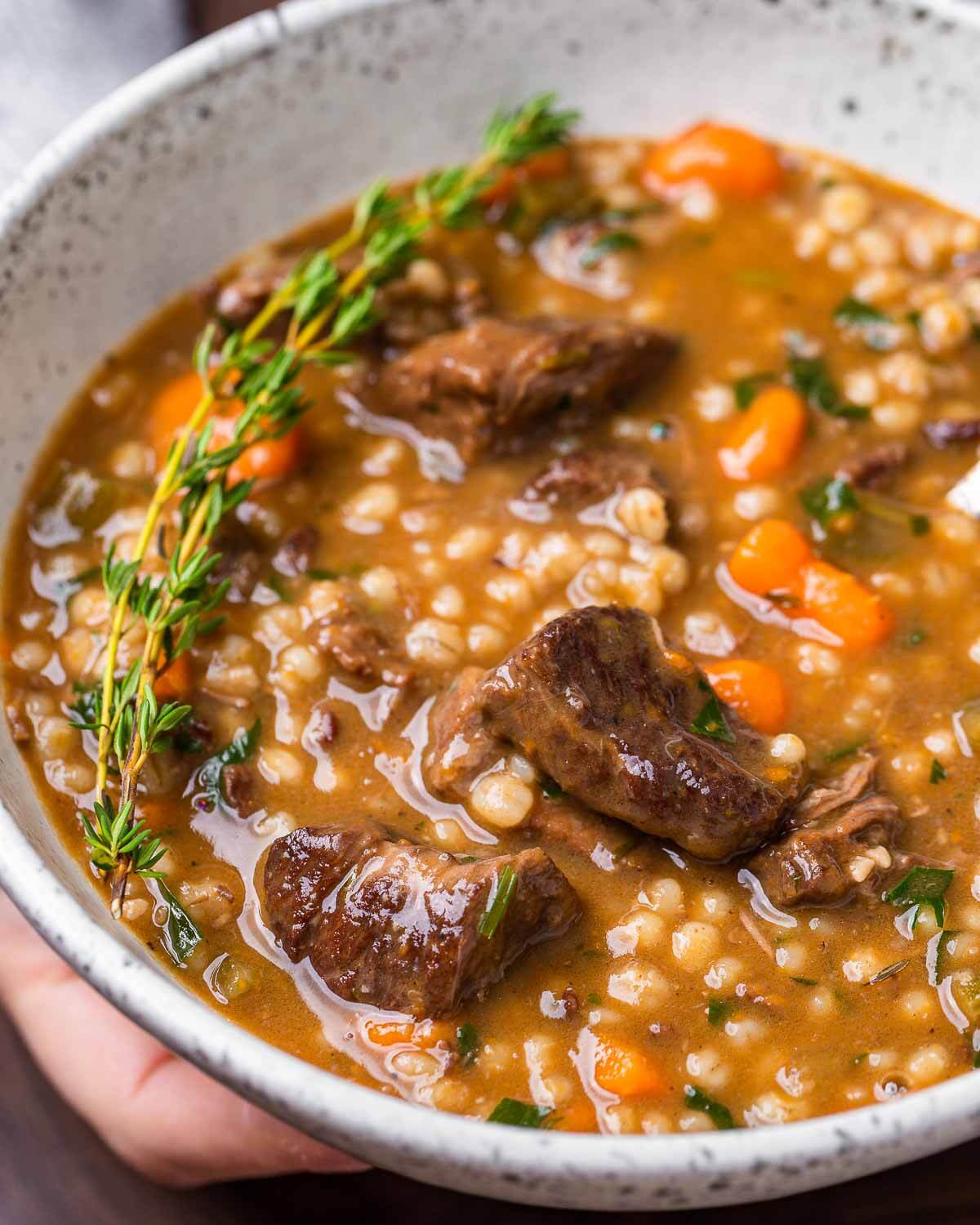 Hands holding large white bowl of beef barley soup.