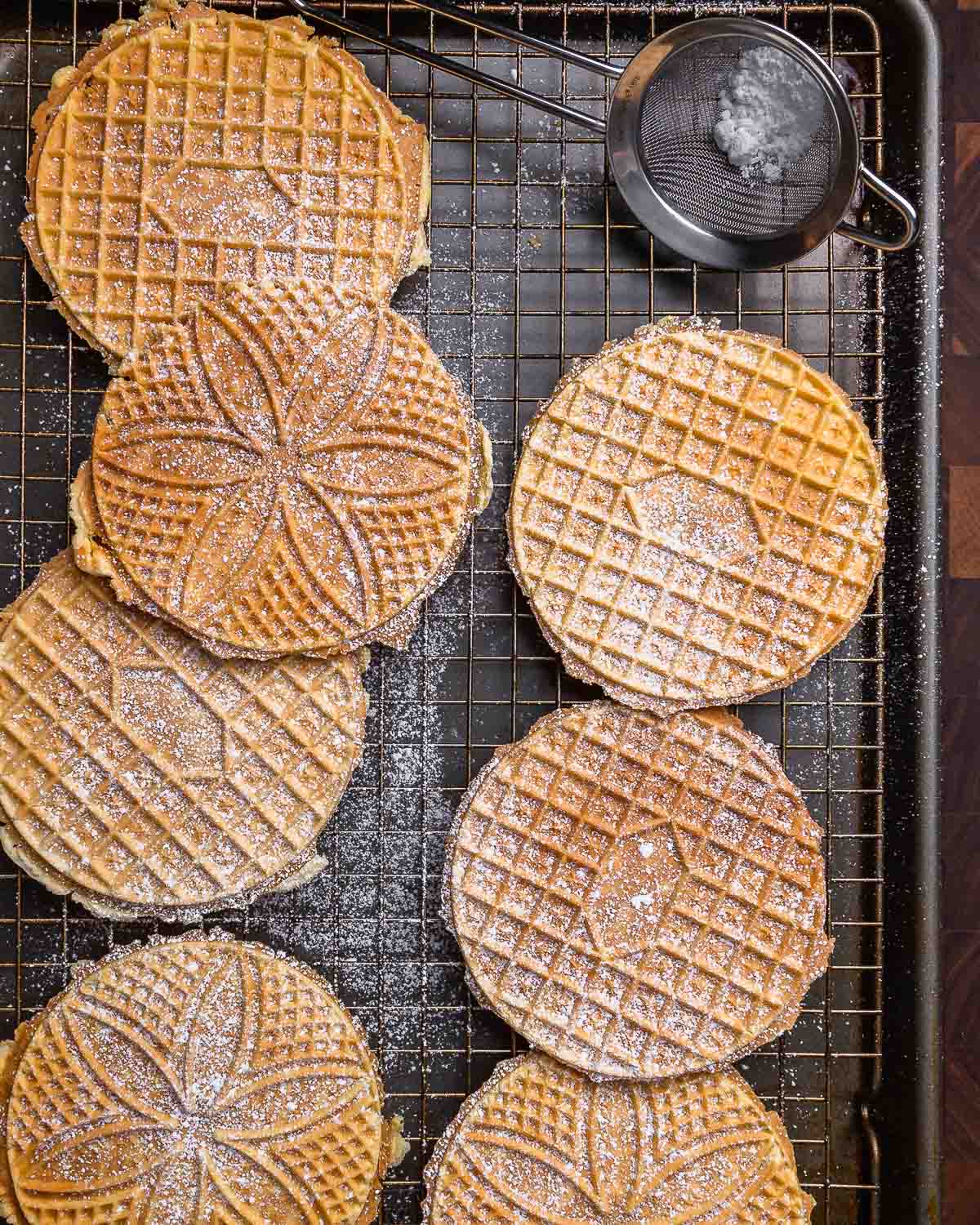 Overhead shot of pizzelles on wire rack with powdered sugar sifter.