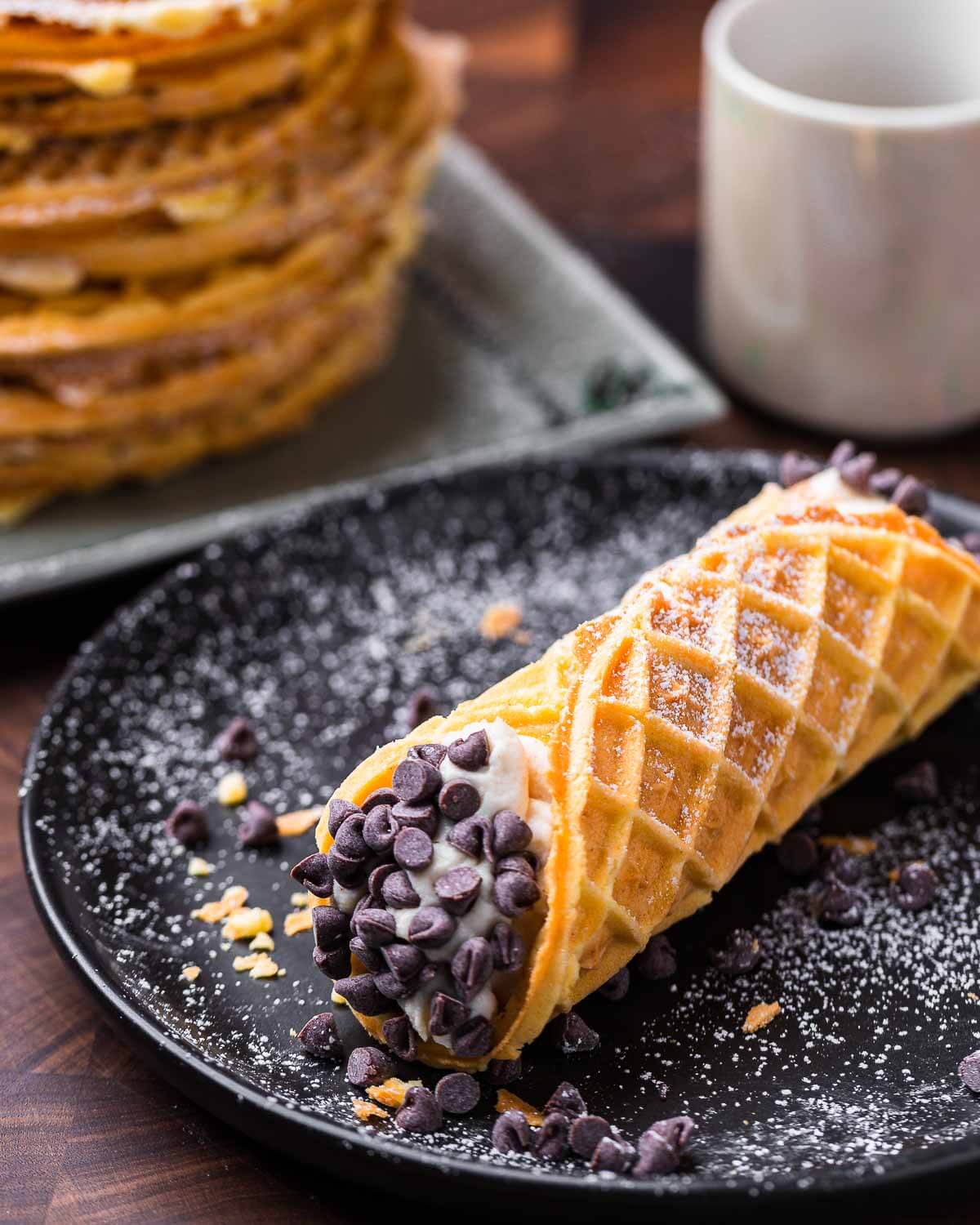 Pizzelle cannoli on black plate with coffee and stack of pizzelles in the background.