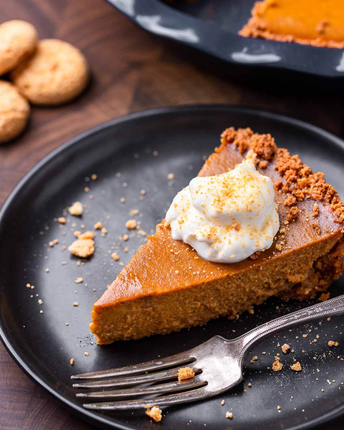 Slice of pumpkin pie with whip cream and crumbled amaretti cookies on black plate.