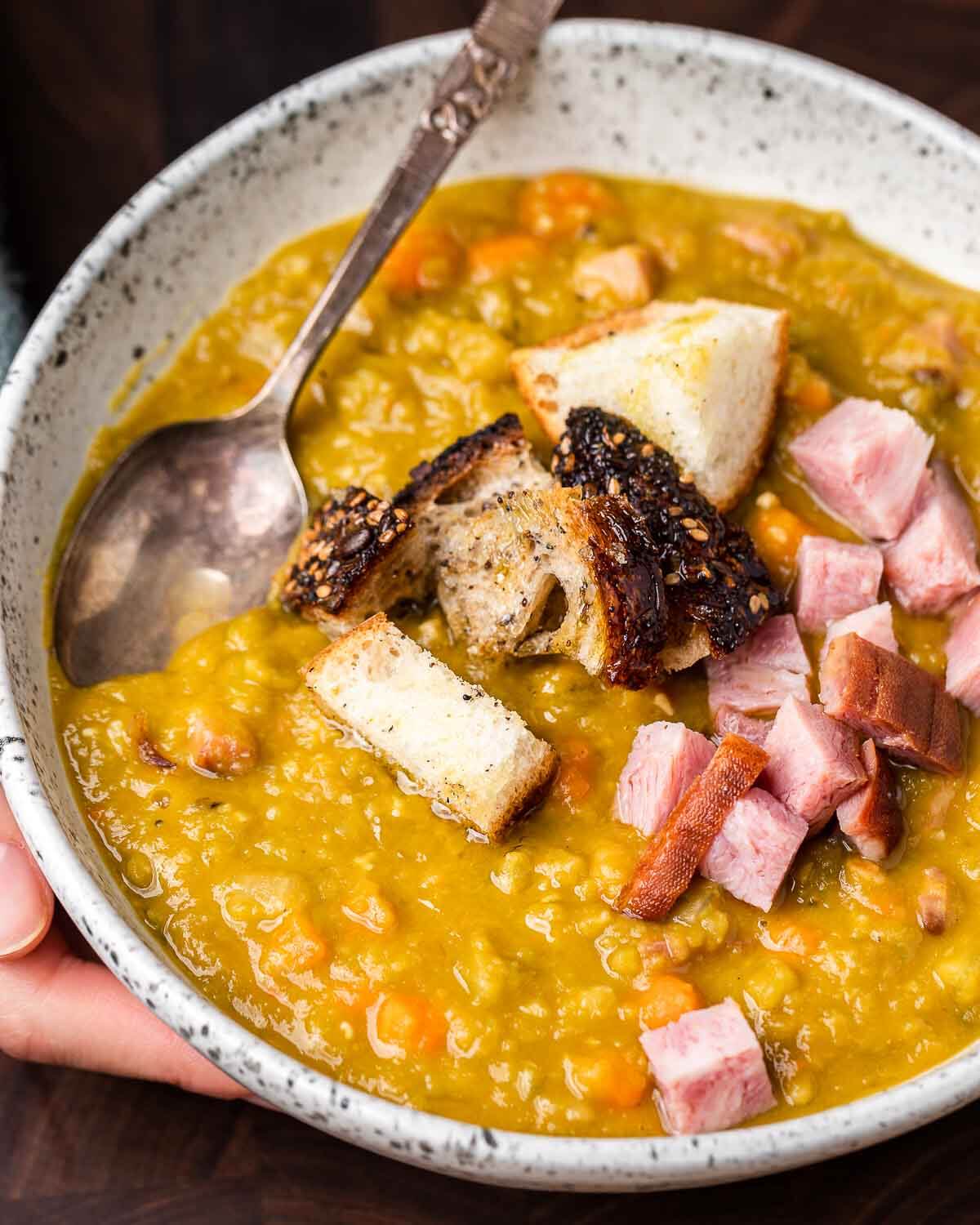 Hands holding white bowl of split pea soup with ham and croutons.