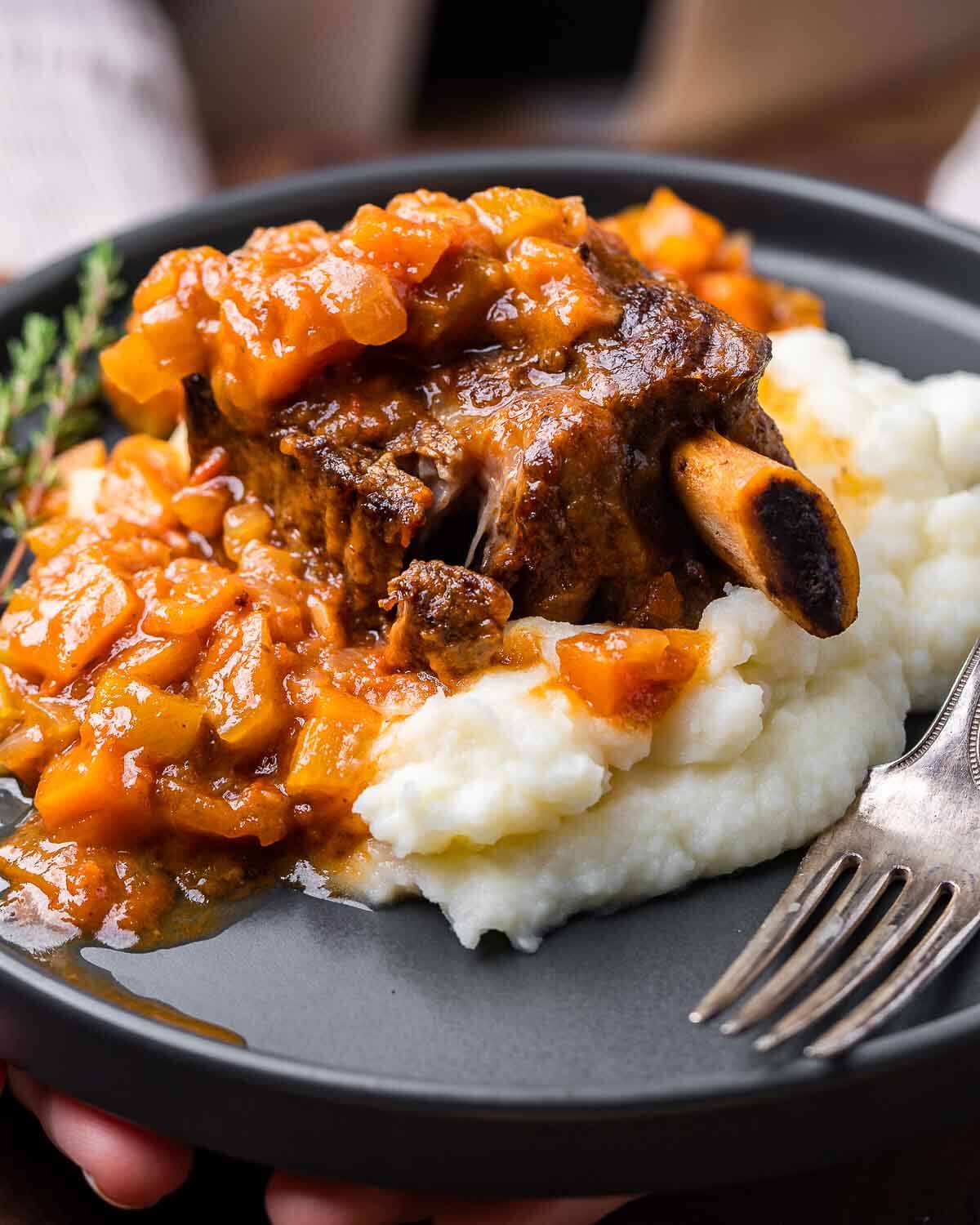 Hands holding plate of braised short ribs on mashed potatoes.