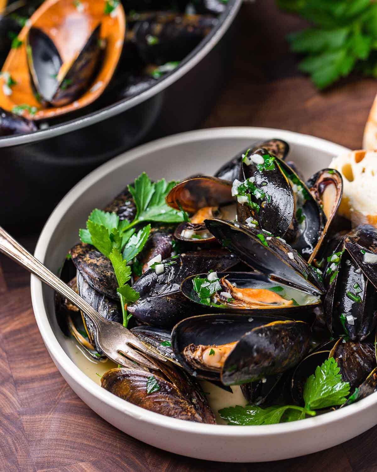 Mussels in white wine sauce in white bowl with tiny fork.