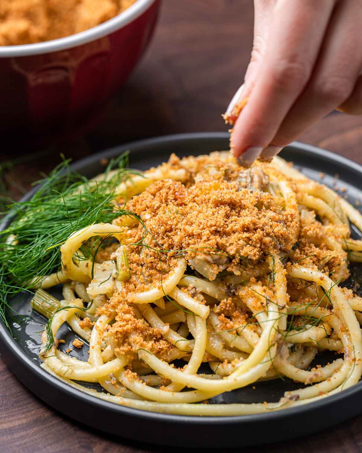 Hands sprinkling breadcrumbs on top of plate of pasta.