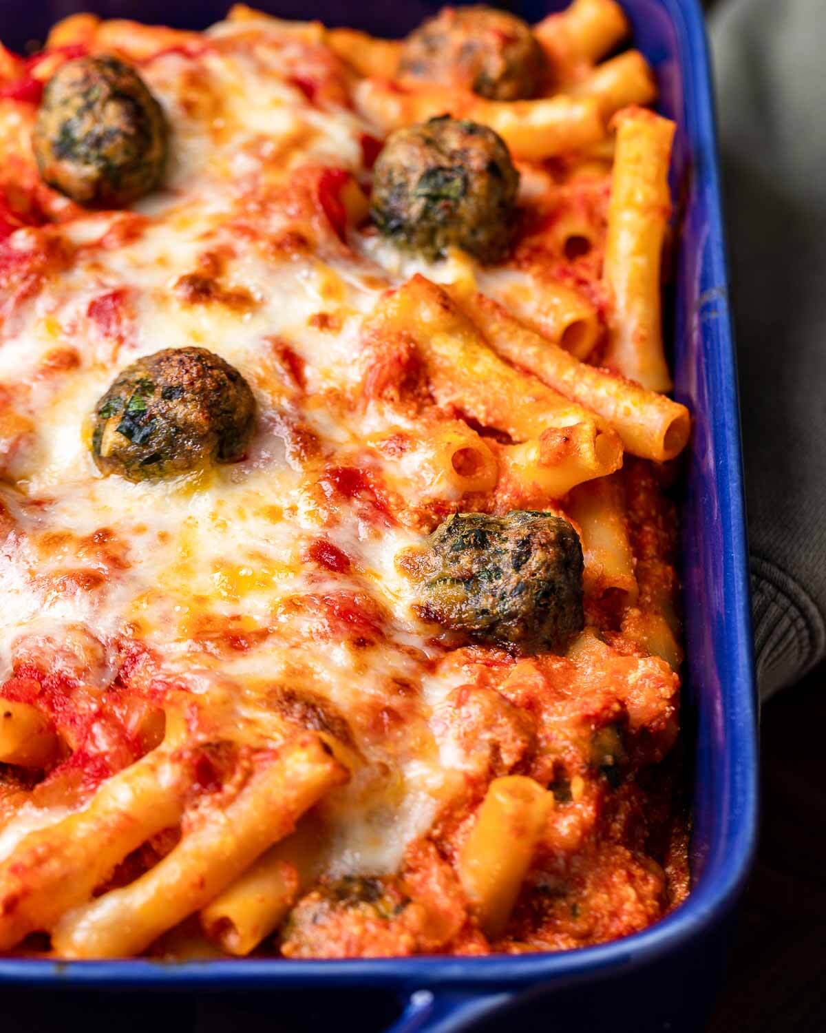 Hands holding blue baking dish with baked ziti and tiny meatballs.