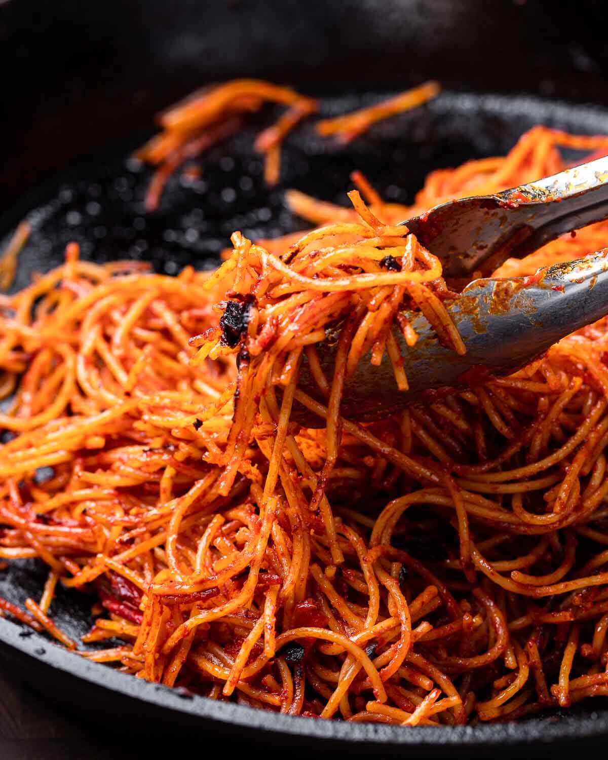 Tongs holding spaghetti all'assassina over cast iron pan.