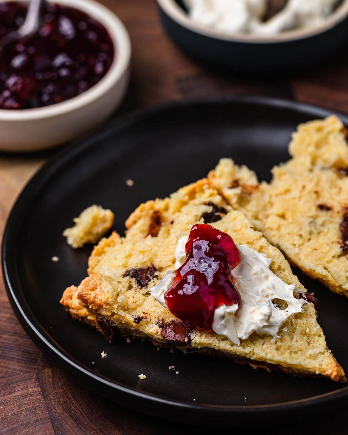 Black plate with chocolate chip scone cut open with jam and clotted cream.