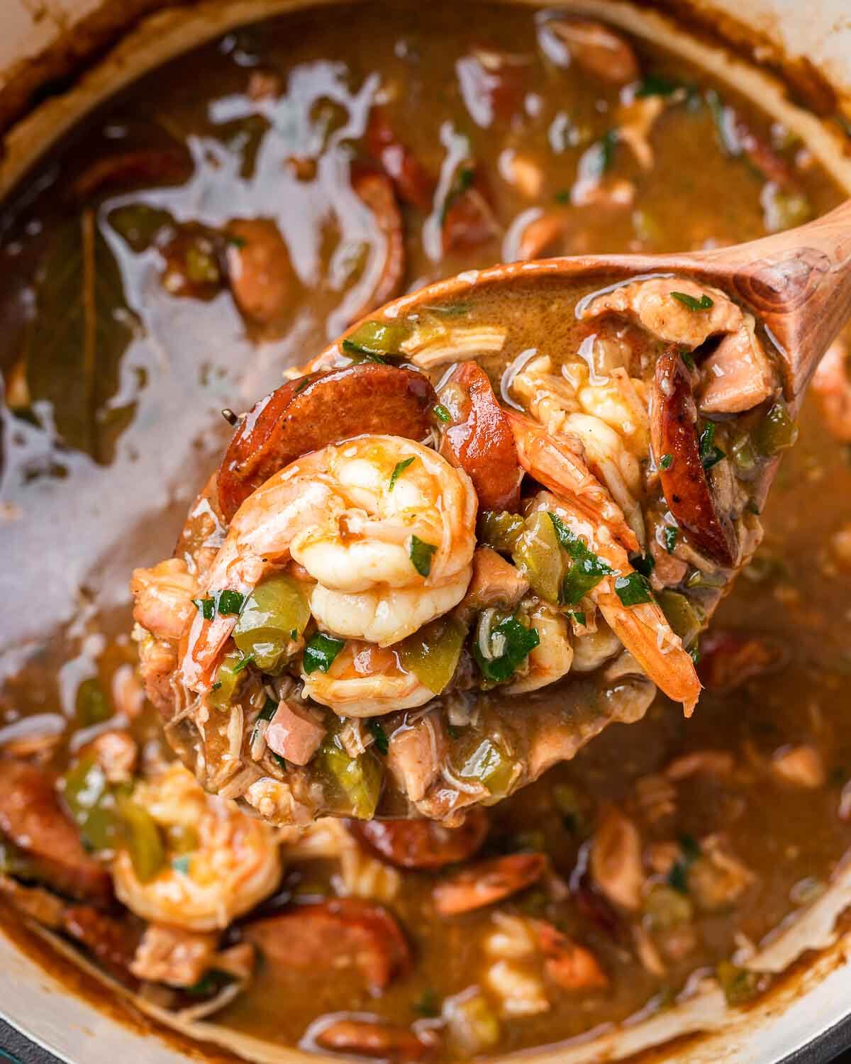 Large wooden spoon holding ladle of chicken, sausage, and shrimp gumbo.