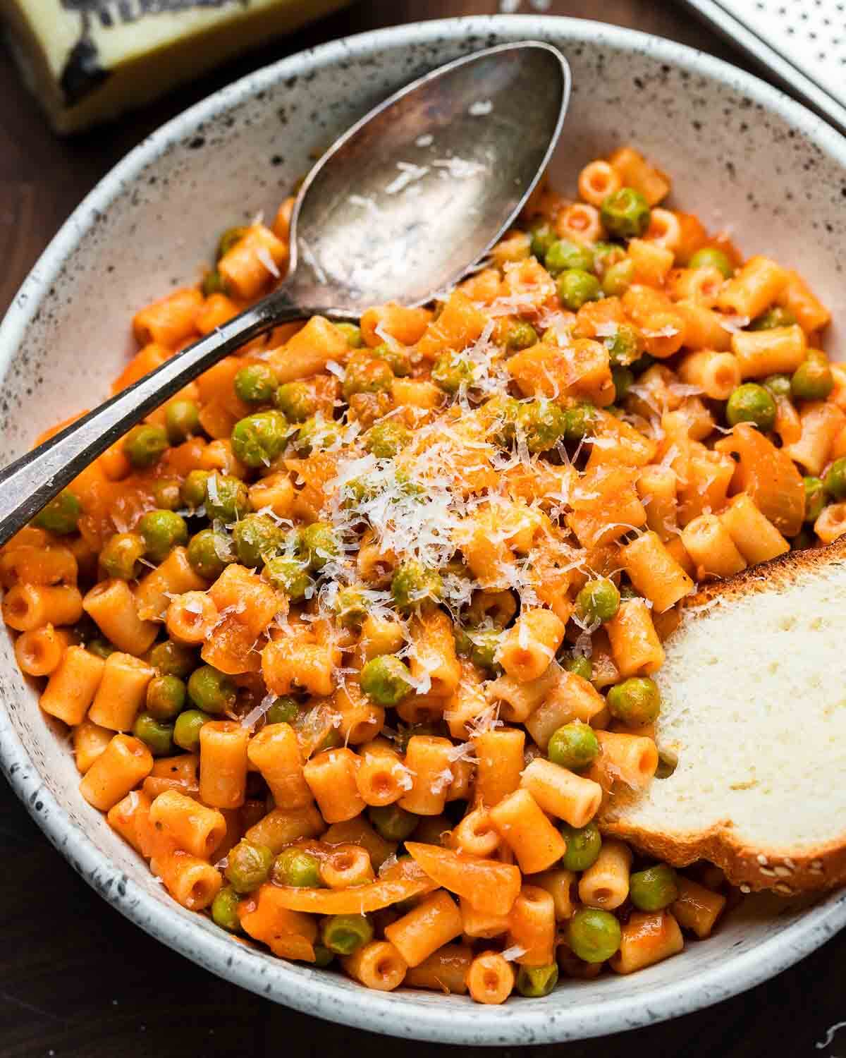 Pasta and peas in red sauce in white bowl with large spoon and piece of bread.