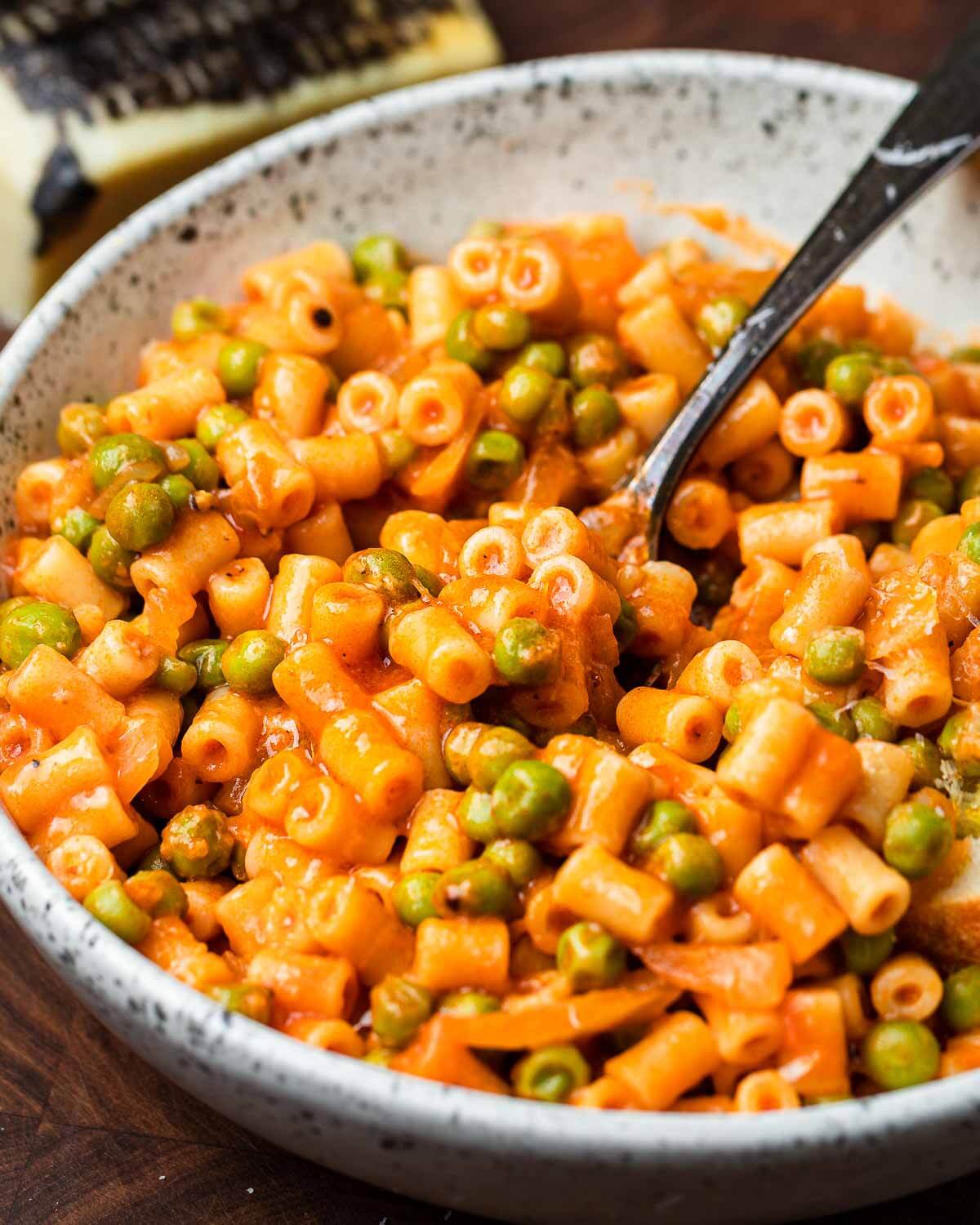Pasta and peas with red sauce in white bowl with block of Pecorino Romano in the background.