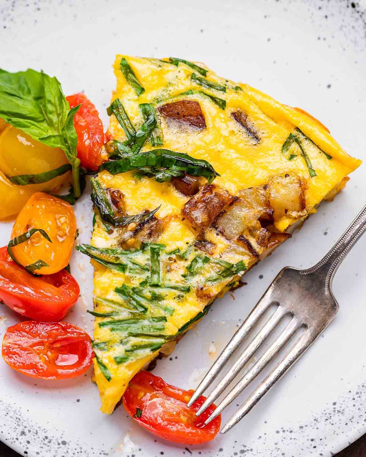 White plate with slice of potato frittata with chives and cherry tomato topping.