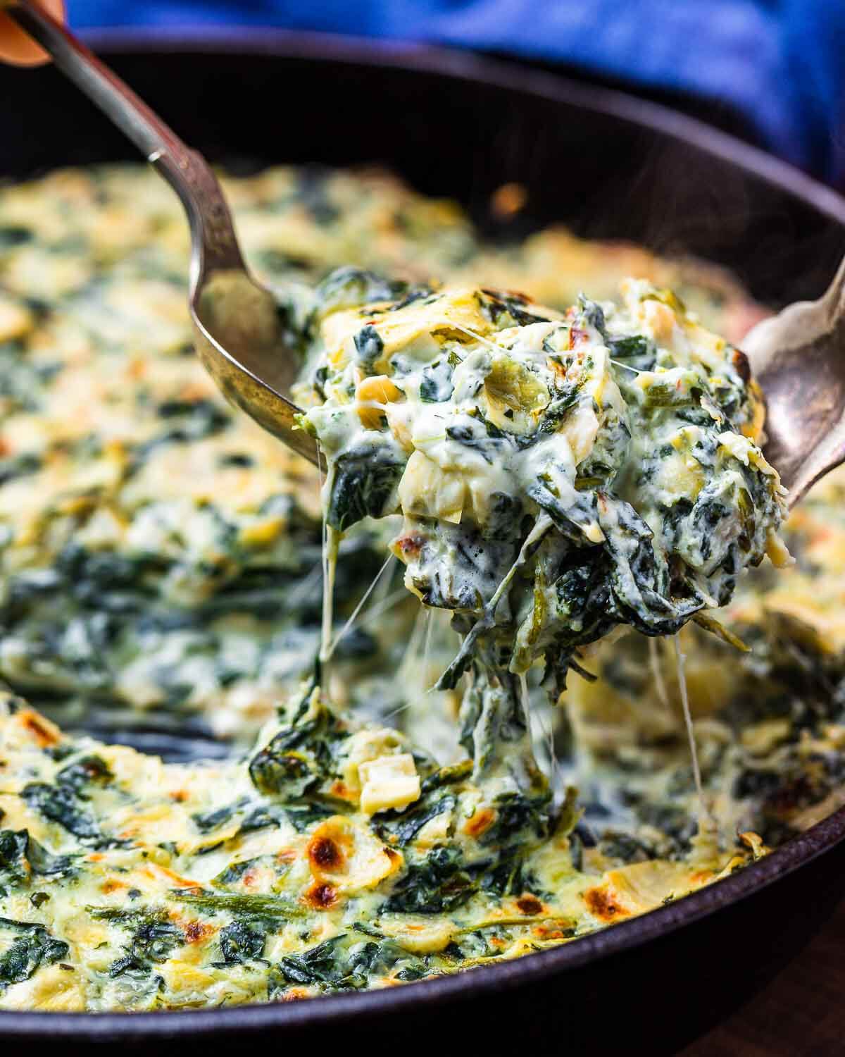 Forks pulling hot spinach artichoke dip out of cast iron pan.