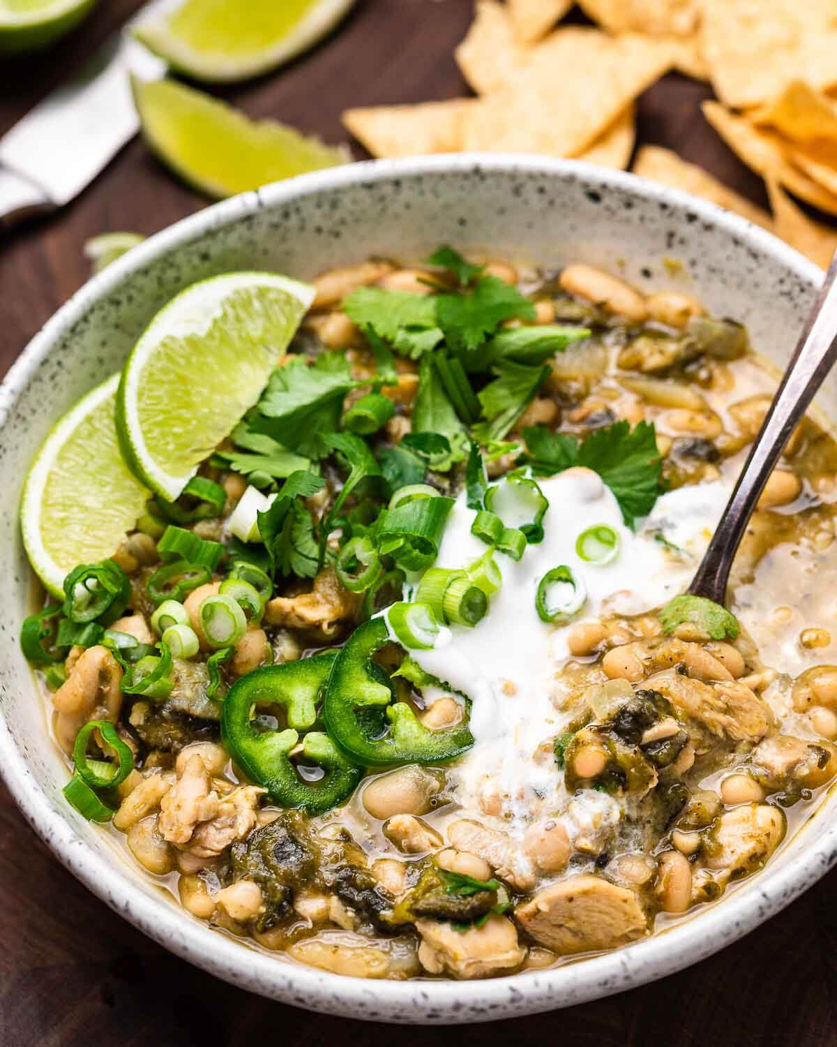 White bowl of white chicken chili topped with limes, green onions, and jalapenos.